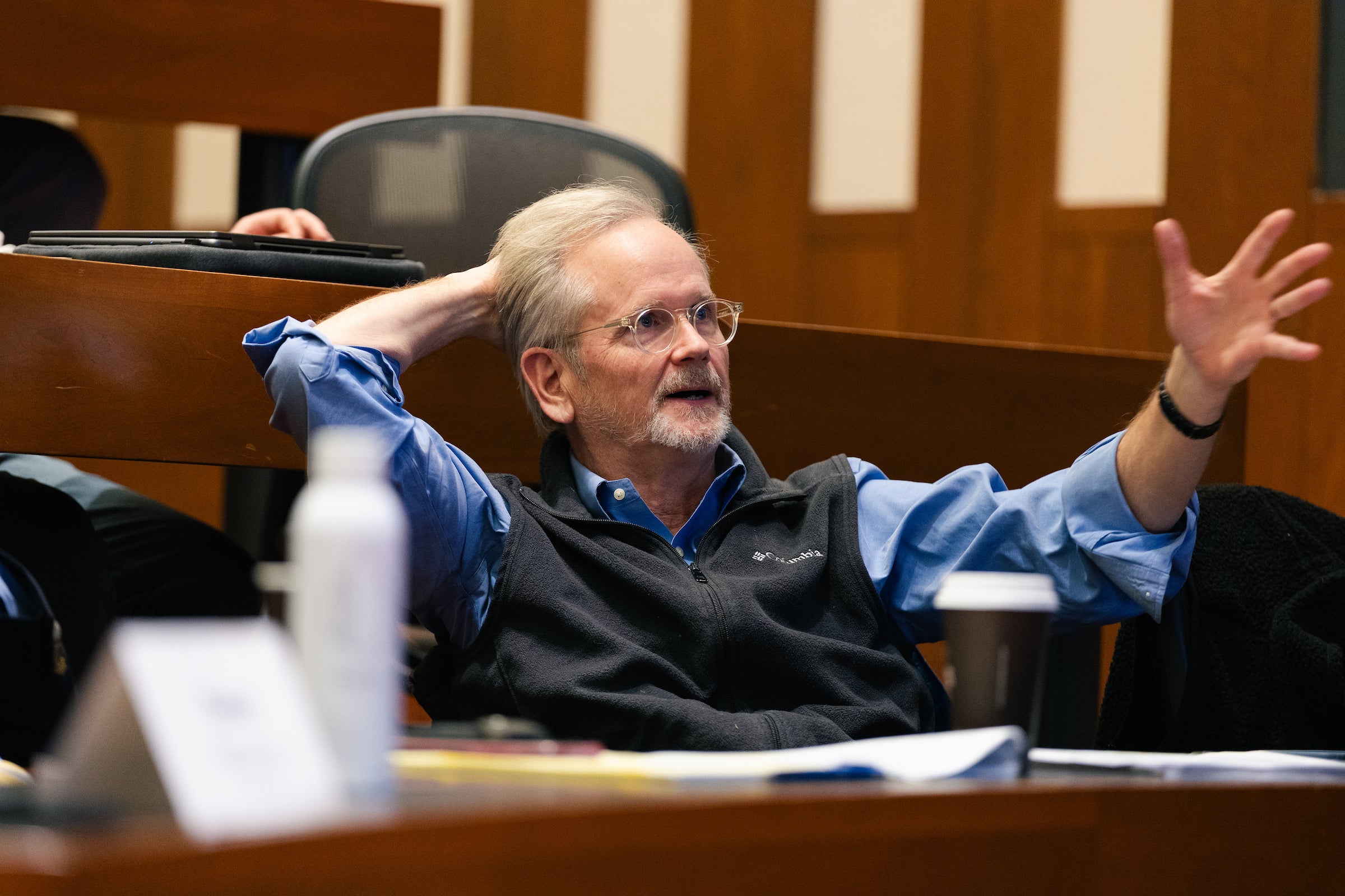 A professor leans back in a chair in a classroom while he makes a comment.