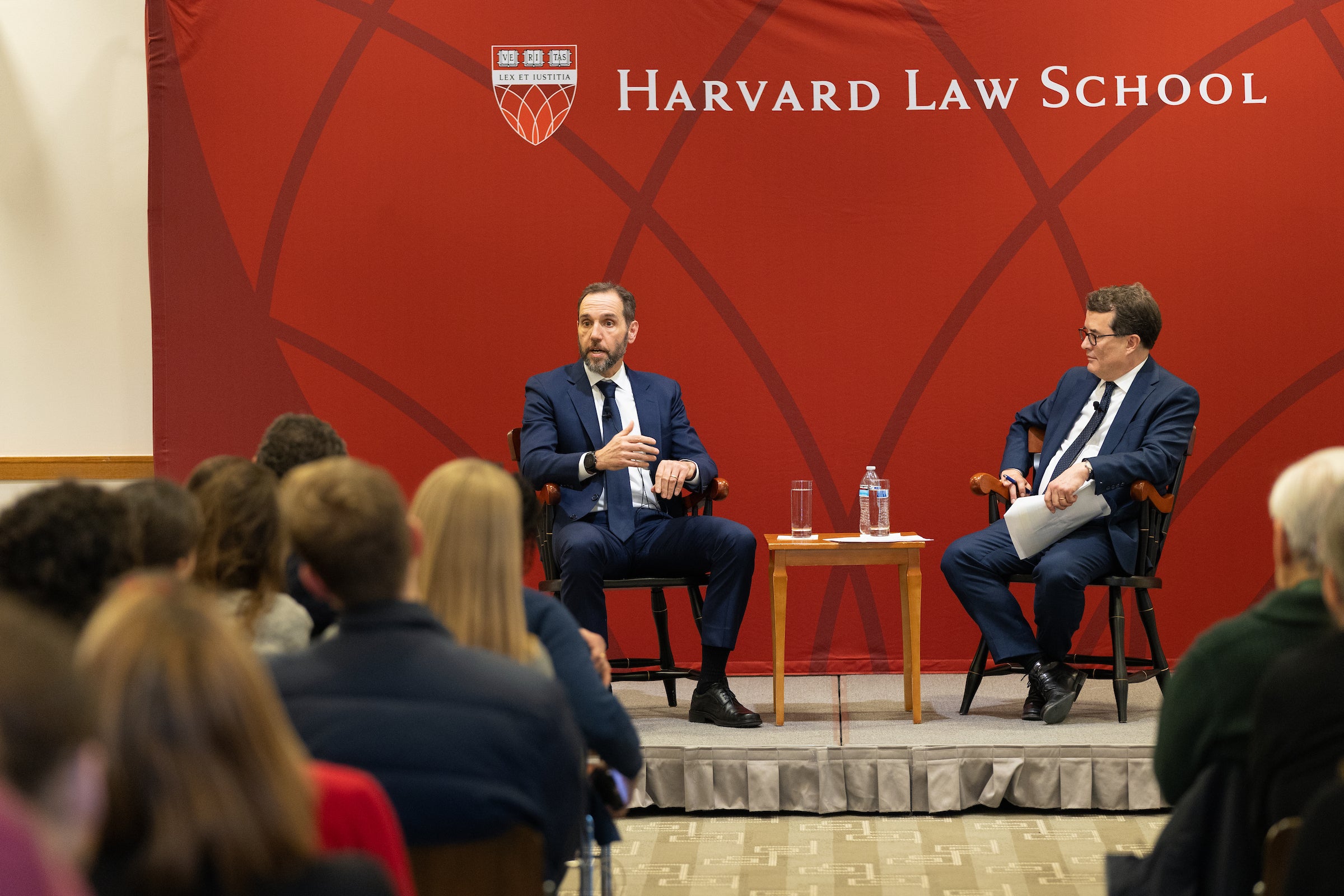Two men sit on a stage in front of an audience.