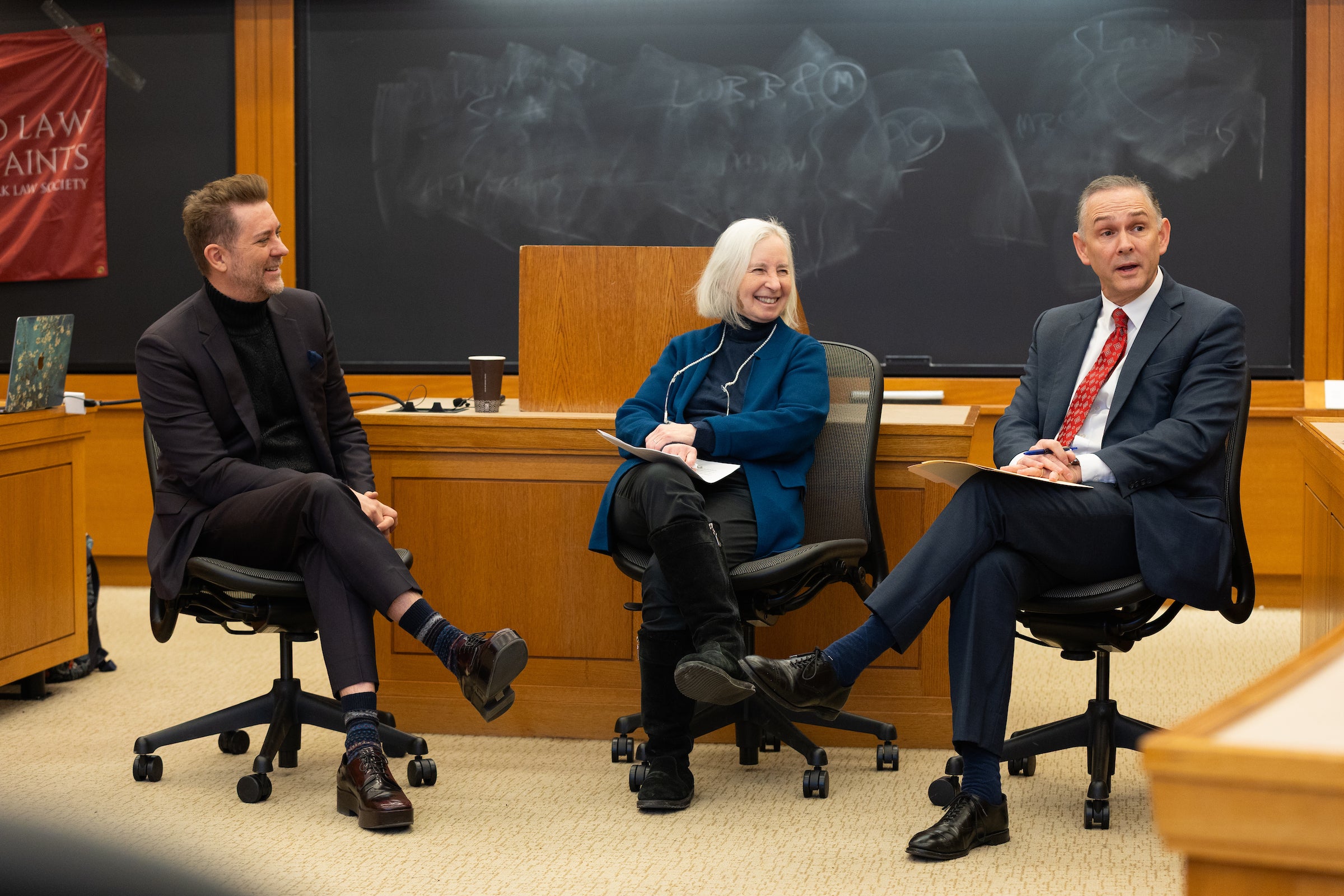 Three people sit in chairs and engage in conversation in a classroom.