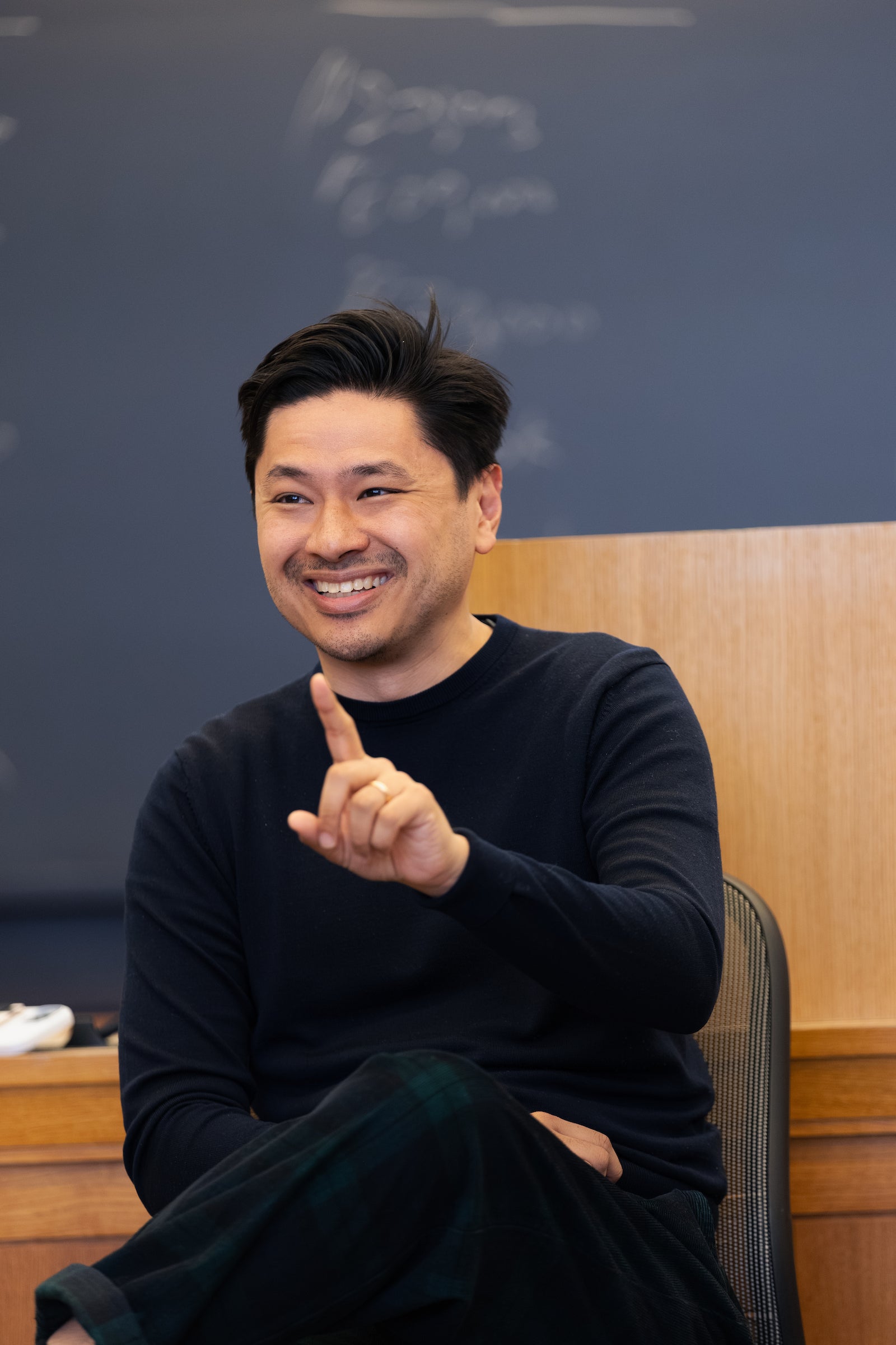 A man sitting in a chair in a classroom speaking to an audience smiles and holds up his index finger.