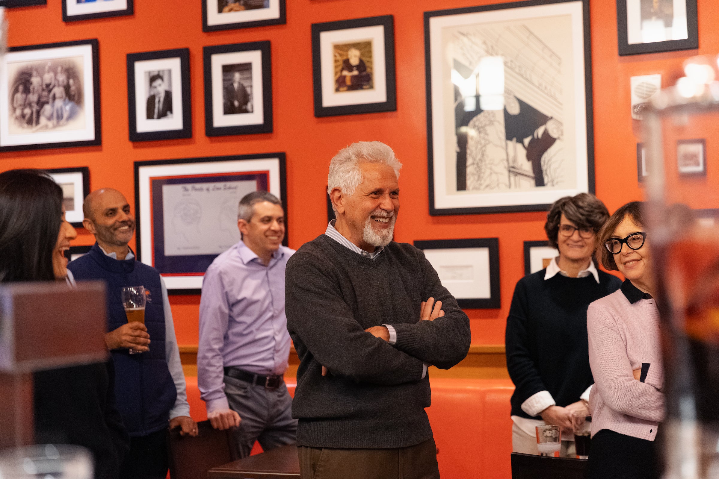 A group of people are gathered around a man in a room with an orange wall adorned with framed photographs, smiling and enjoying drinks together.