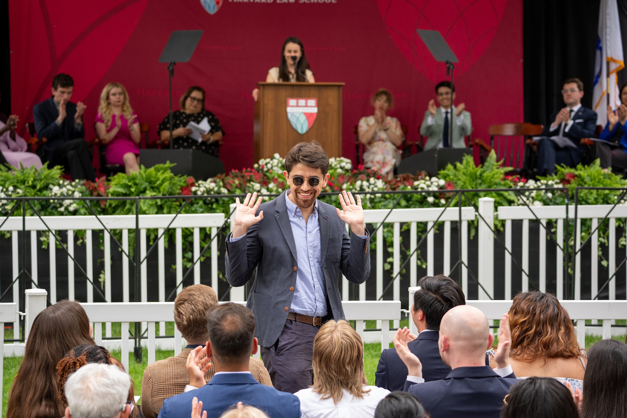 A person in a suit and sunglasses waves to an audience at an outdoor ceremony, with a speaker and panel seated behind them.
