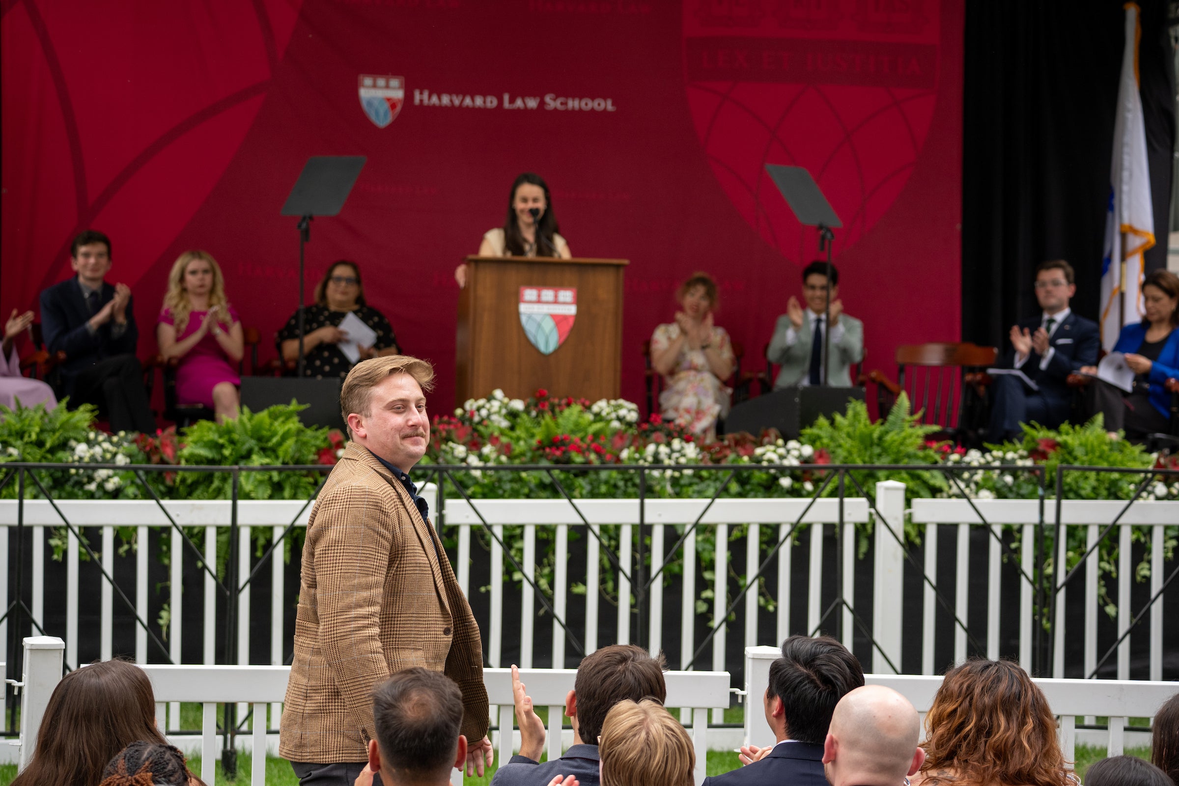 A person in a plaid jacket stands in the foreground at an outdoor event as he is recognized for an award, with a speaker addressing an audience from a podium on a stage behind him.