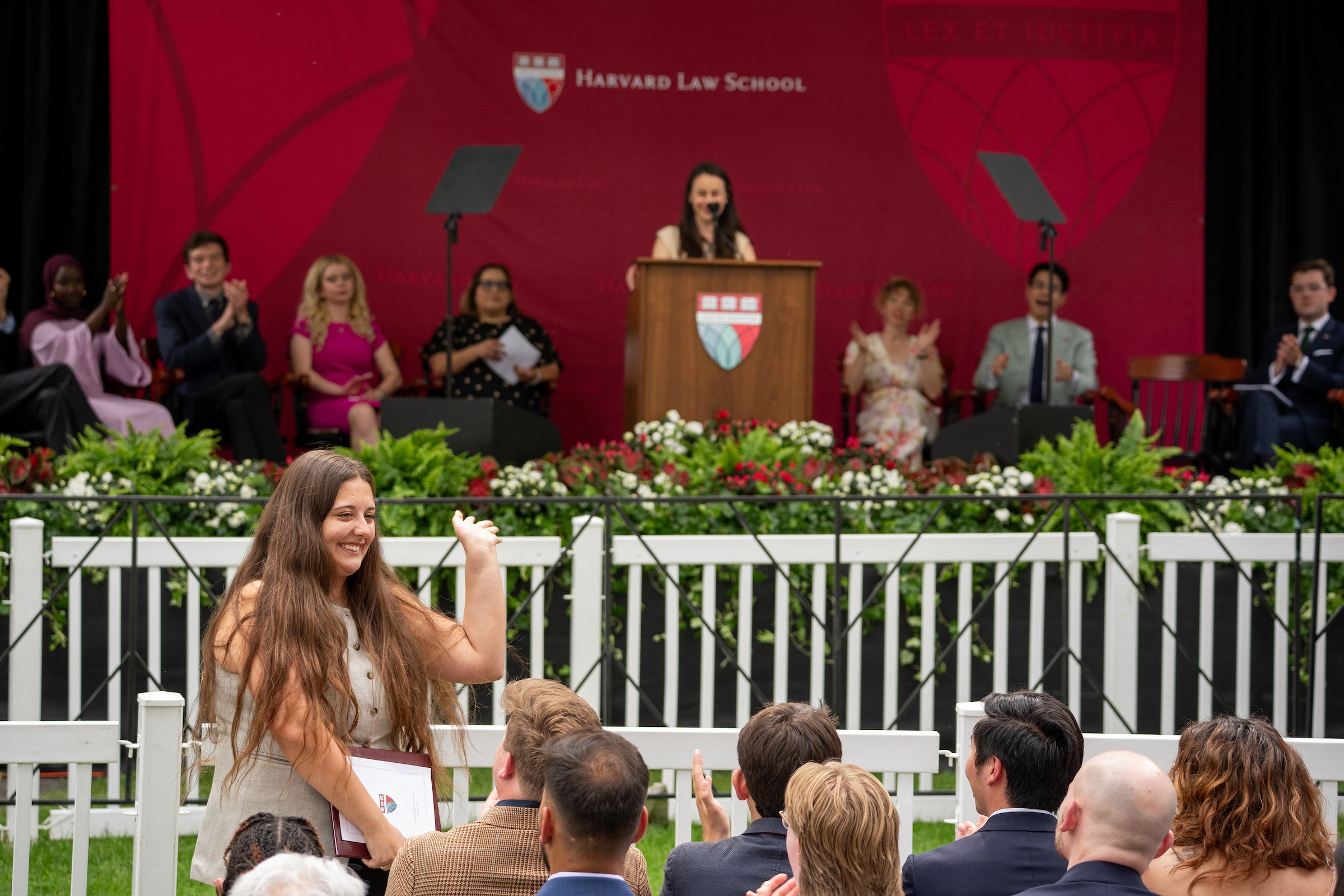 A graduate smiles and waves from the audience as she is recognized for an award, with a speaker and applauding guests on stage behind her.