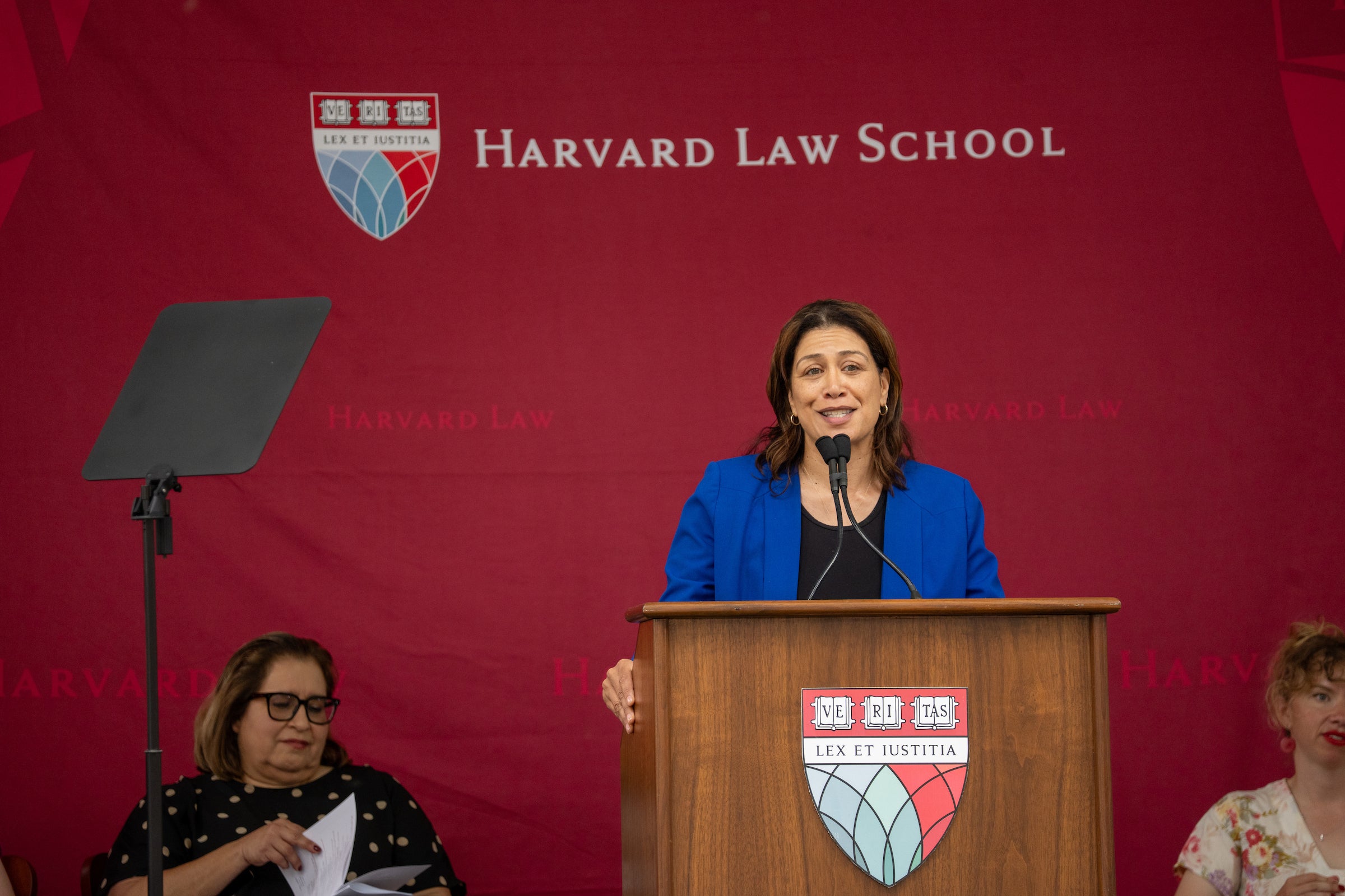 A person in a blue jacket stands at a wooden podium with the Harvard Law School logo, speaking into a microphone during an event, with two other individuals seated nearby.
