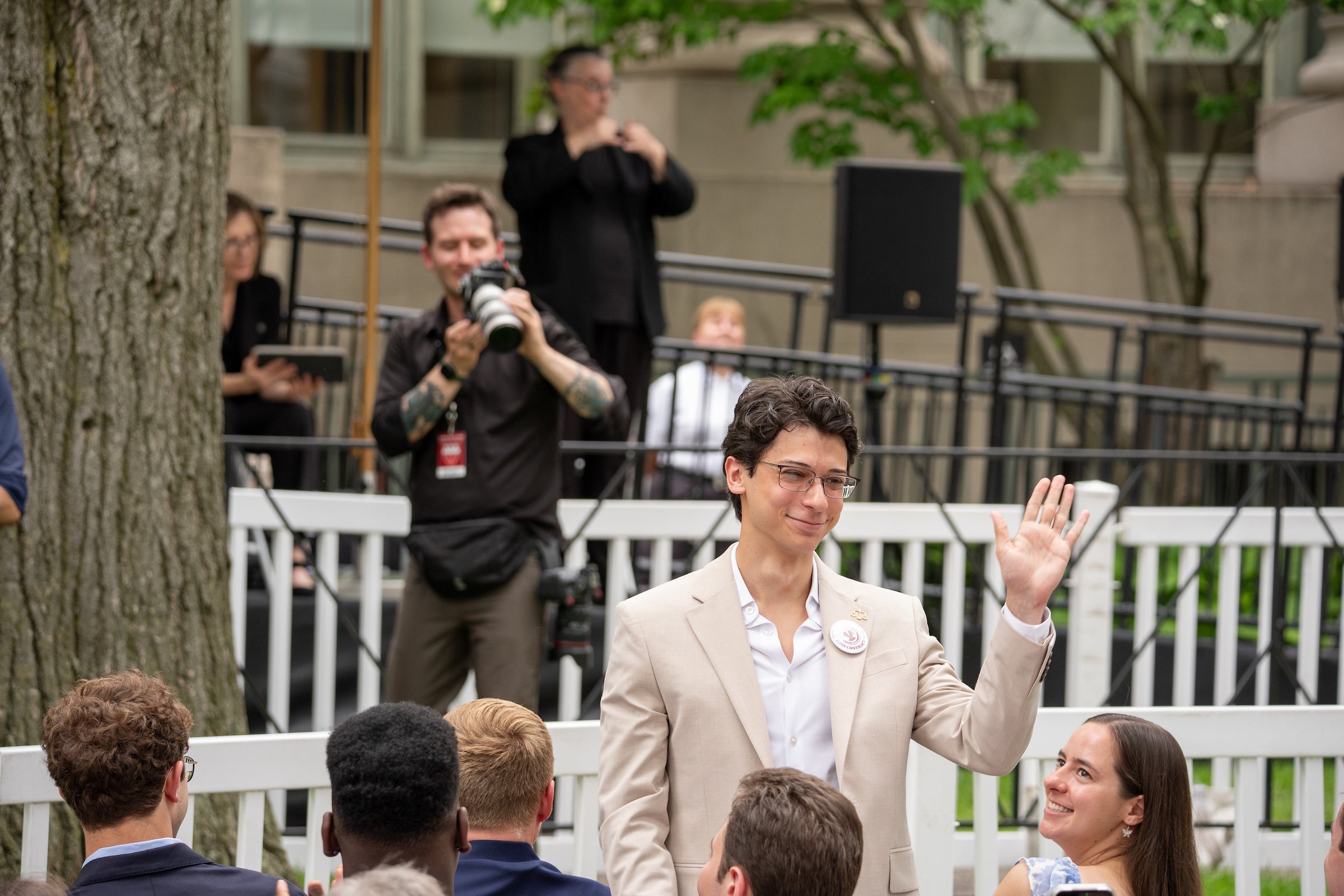 A person in a light-colored suit waves to an audience while standing outdoors near a white fence.