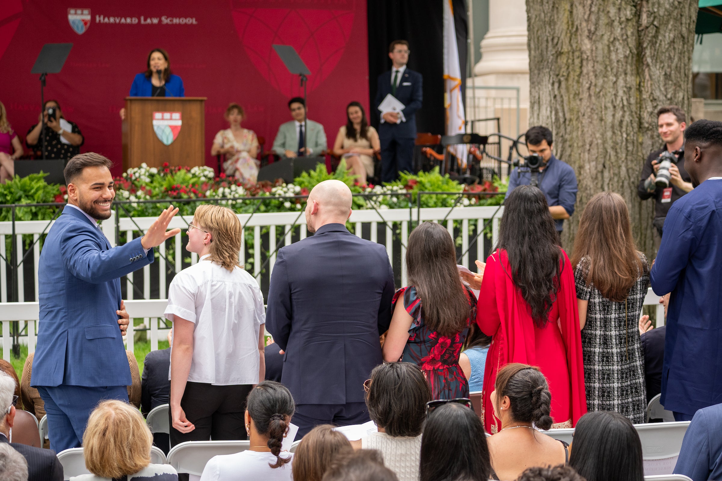A person in a blue suit waves to an audience at an outdoor event hosted by Harvard Law School, with speakers and photographers in the background.