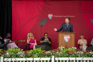 A person speaks at a podium in front of several seated attendees, with a floral arrangement in the foreground.