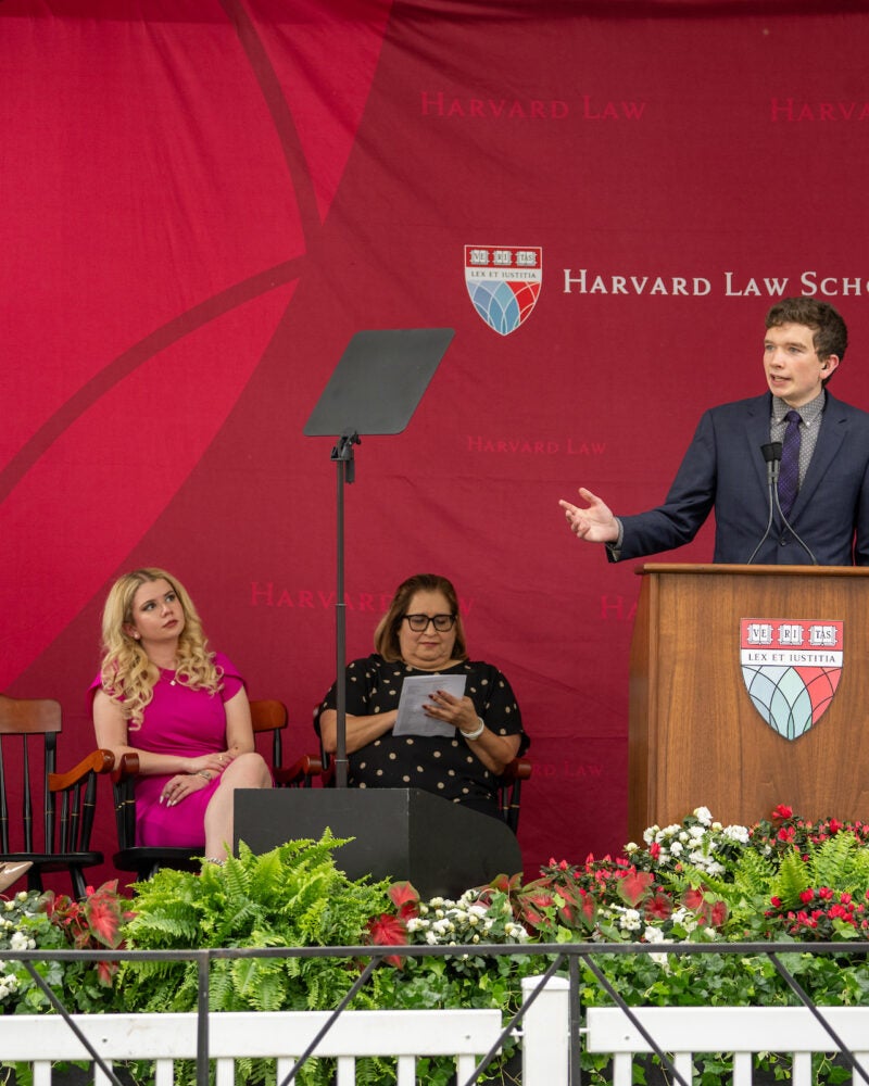 A person speaks at a podium in front of several seated attendees, with a floral arrangement in the foreground.