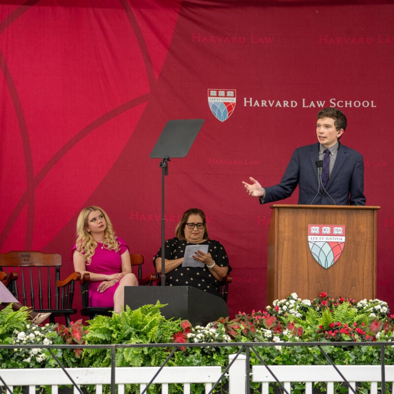 A person speaks at a podium in front of several seated attendees, with a floral arrangement in the foreground.
