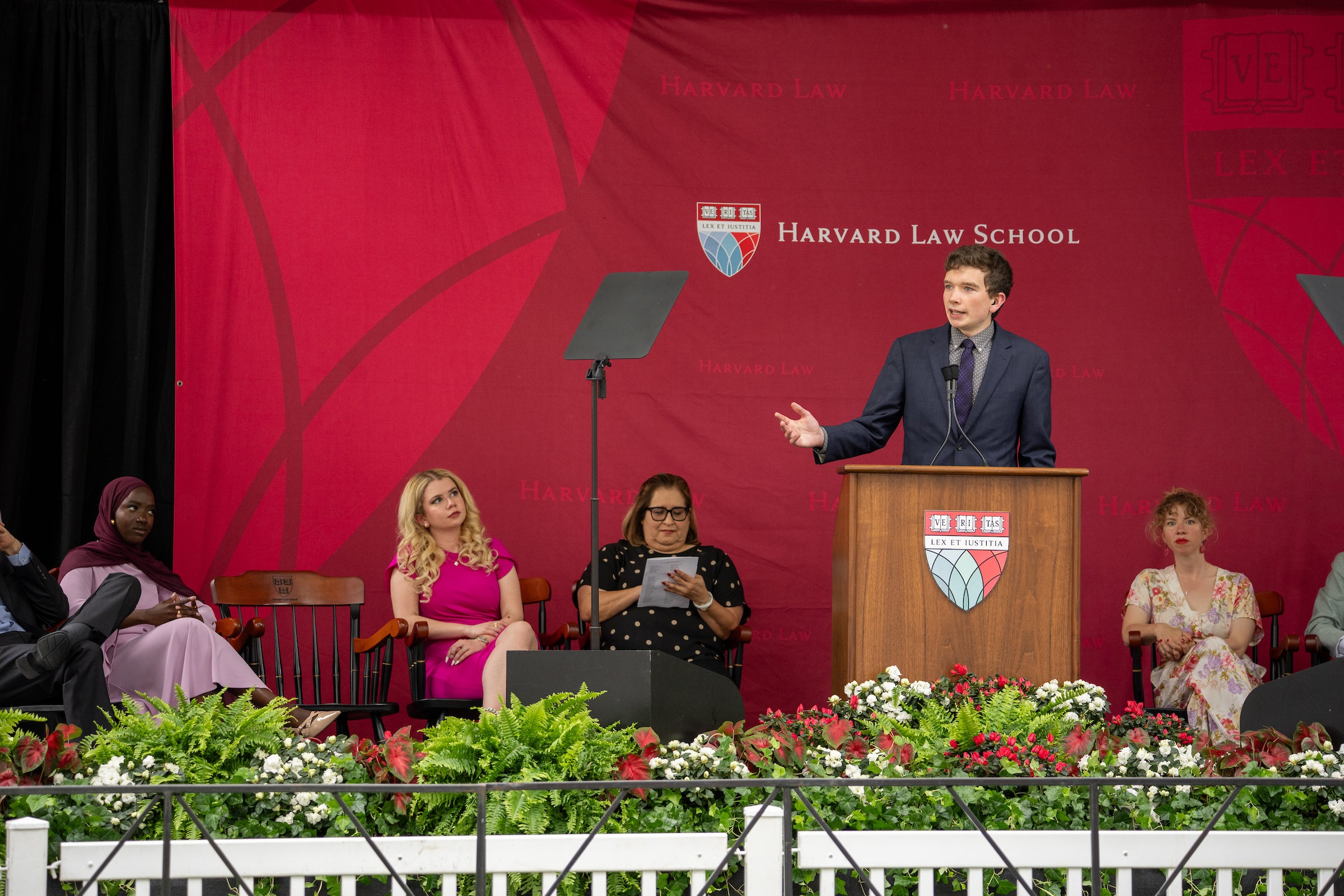 A person speaks at a podium in front of several seated attendees, with a floral arrangement in the foreground.