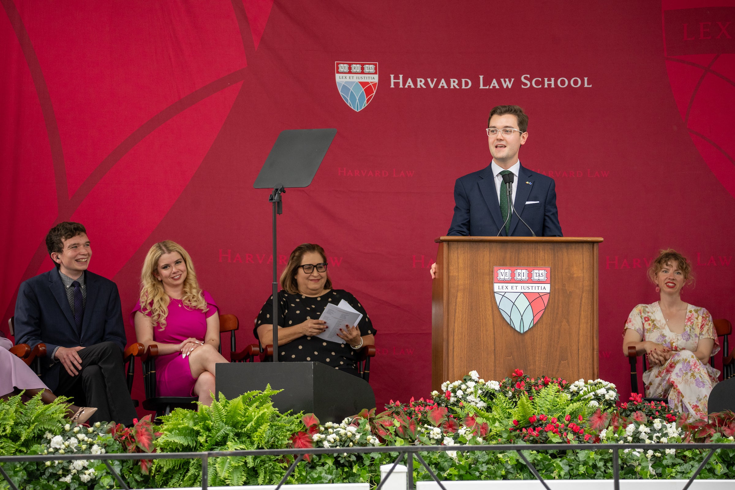 A person speaks at a podium, with several others seated nearby on a stage adorned with floral arrangements.