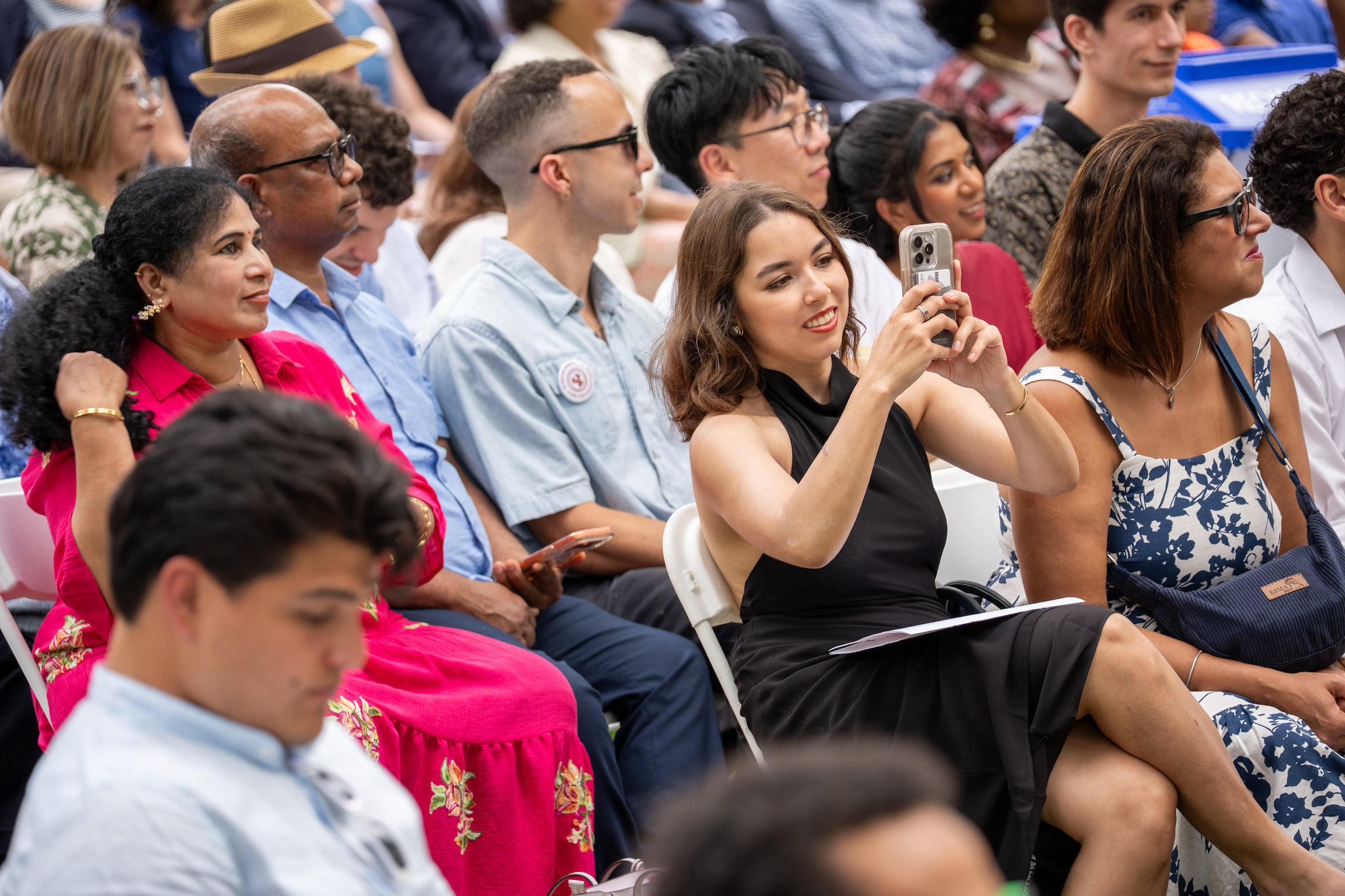 A person in a black dress sits among an audience, holding a phone to take a photo while others watch attentively.