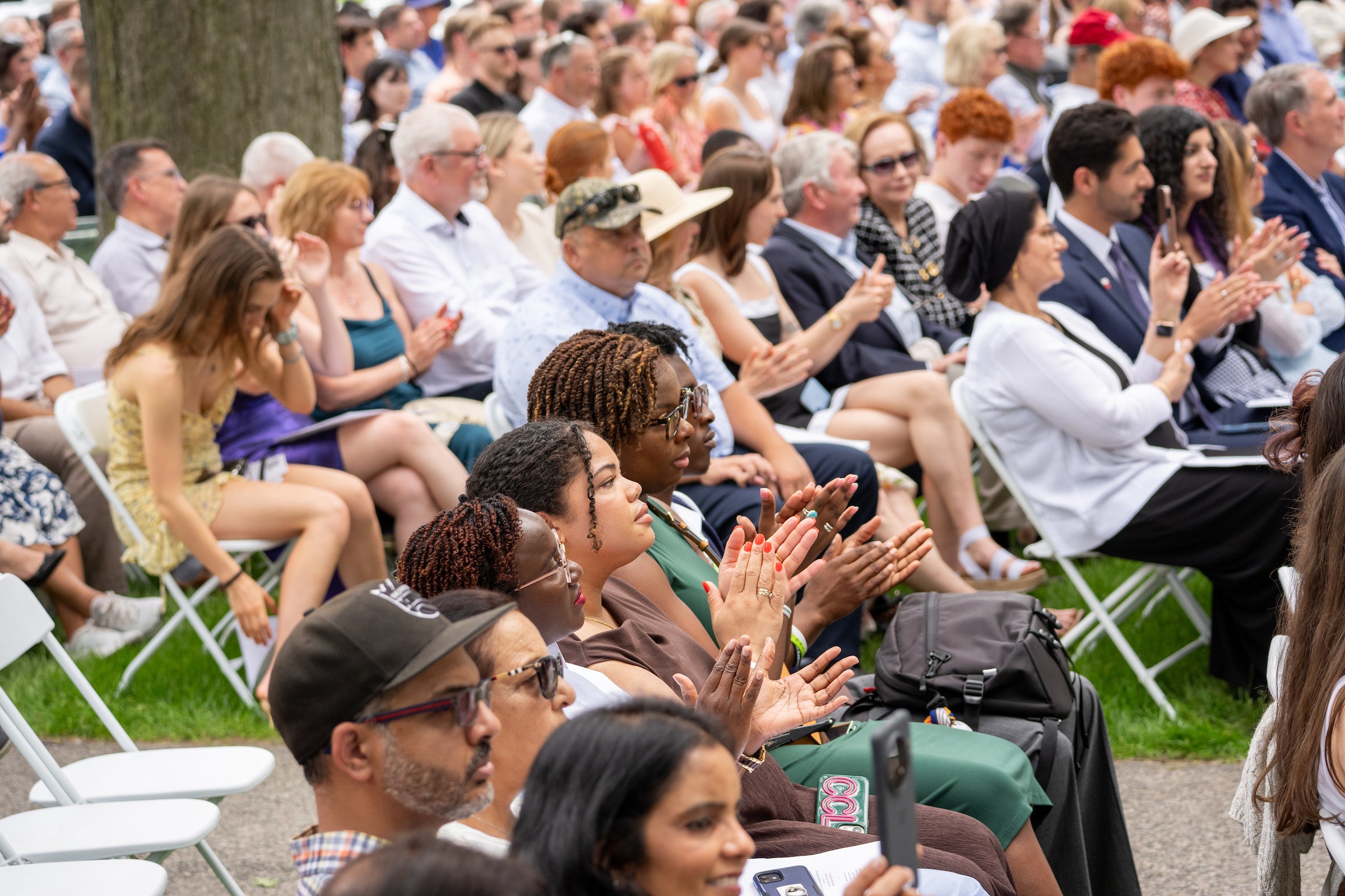 An audience sits outdoors in folding chairs, clapping their hands during an event.