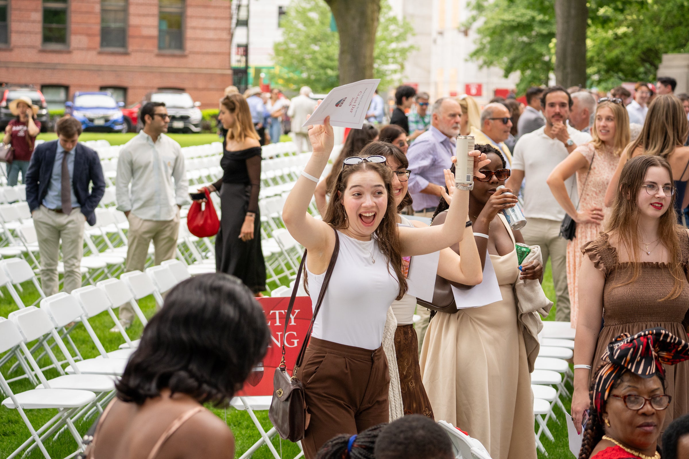 A joyful person in a white top and brown pants raises their arms, holding an event program and a can, amidst a crowd at an outdoor event with rows of white chairs on green grass.