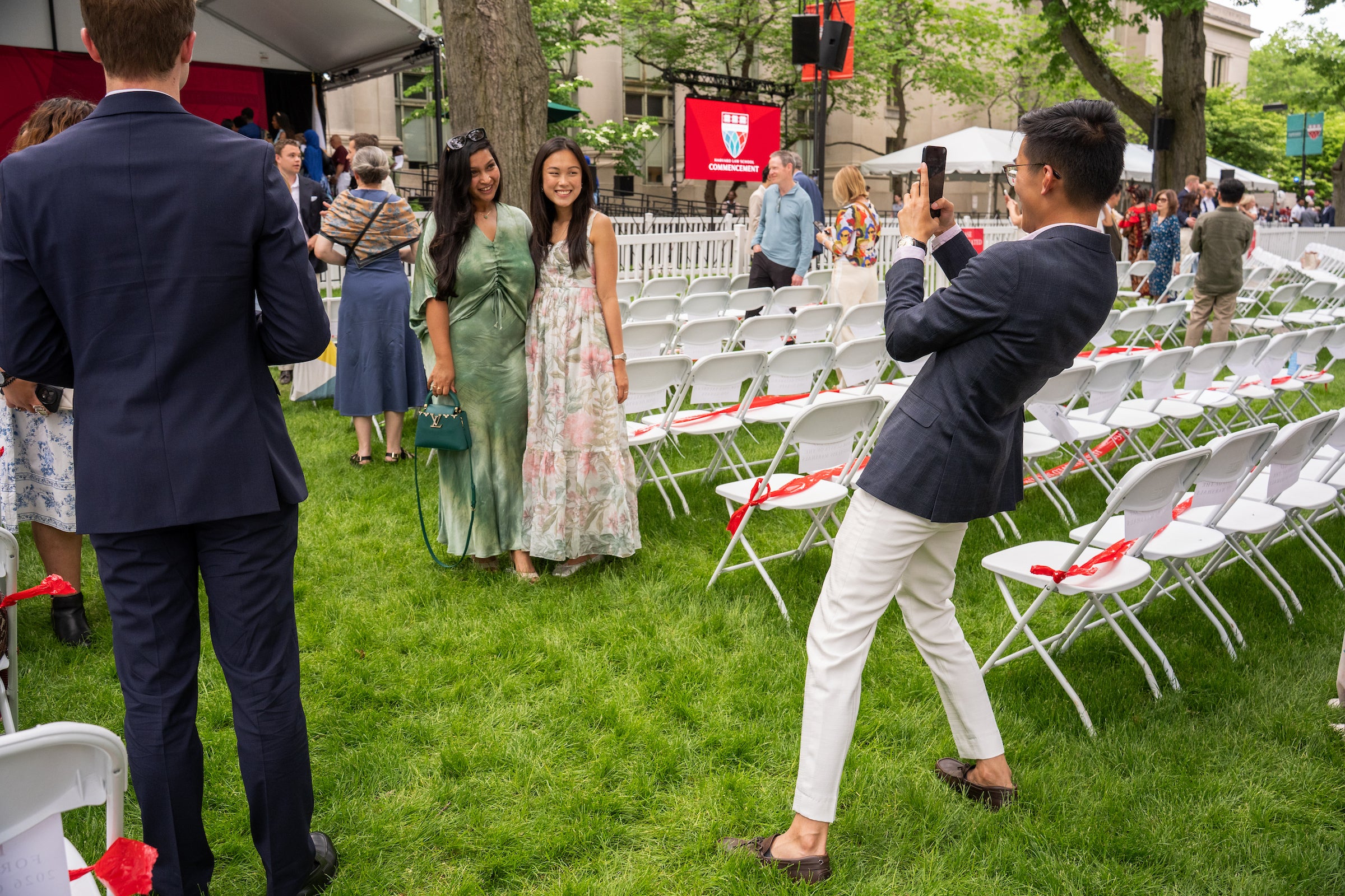 A person takes a photo of two individuals smiling on a grassy field with rows of white chairs, at an outdoor event.