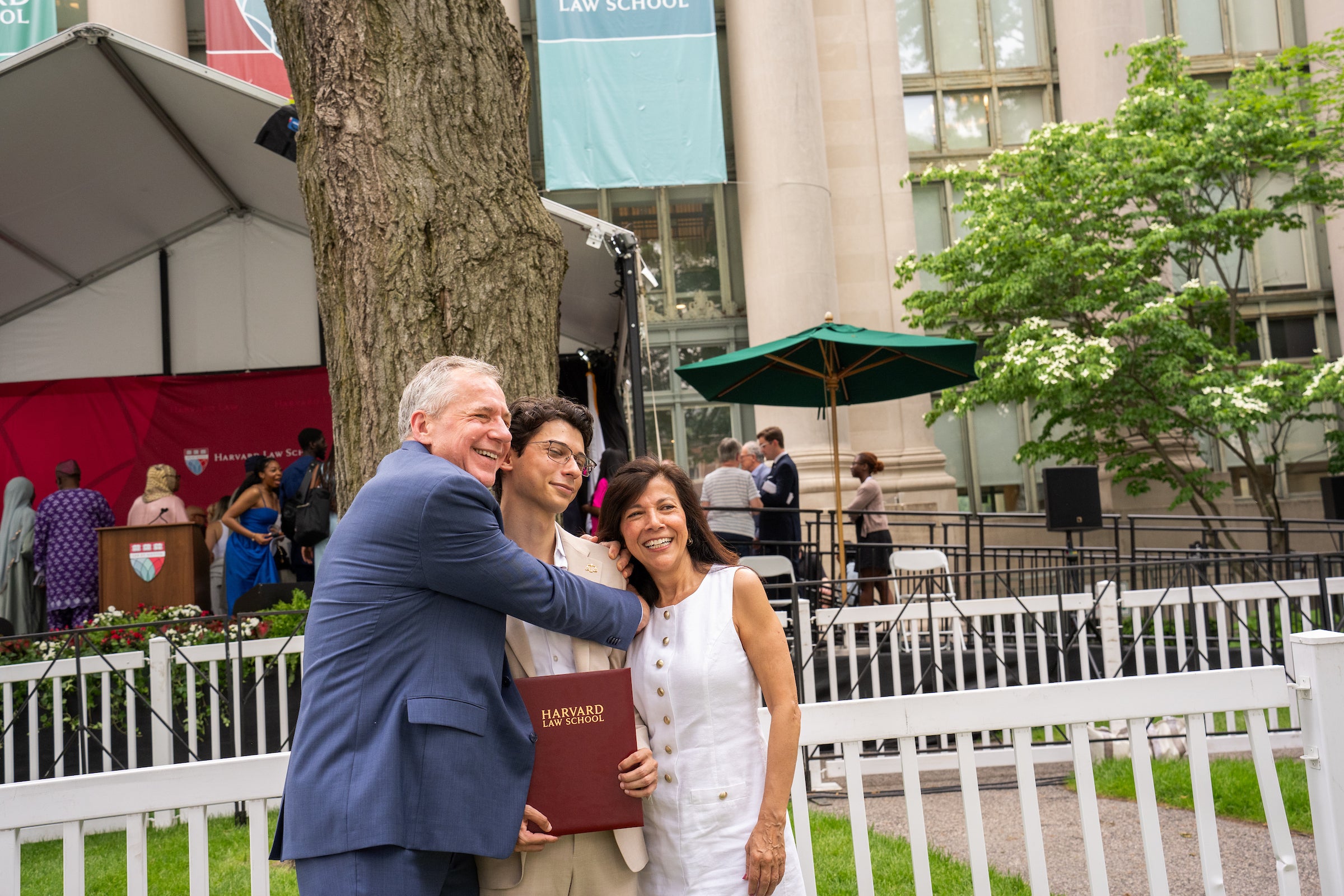 Three people pose happily outdoors, with one holding a Harvard Law School award, in front of the stage.