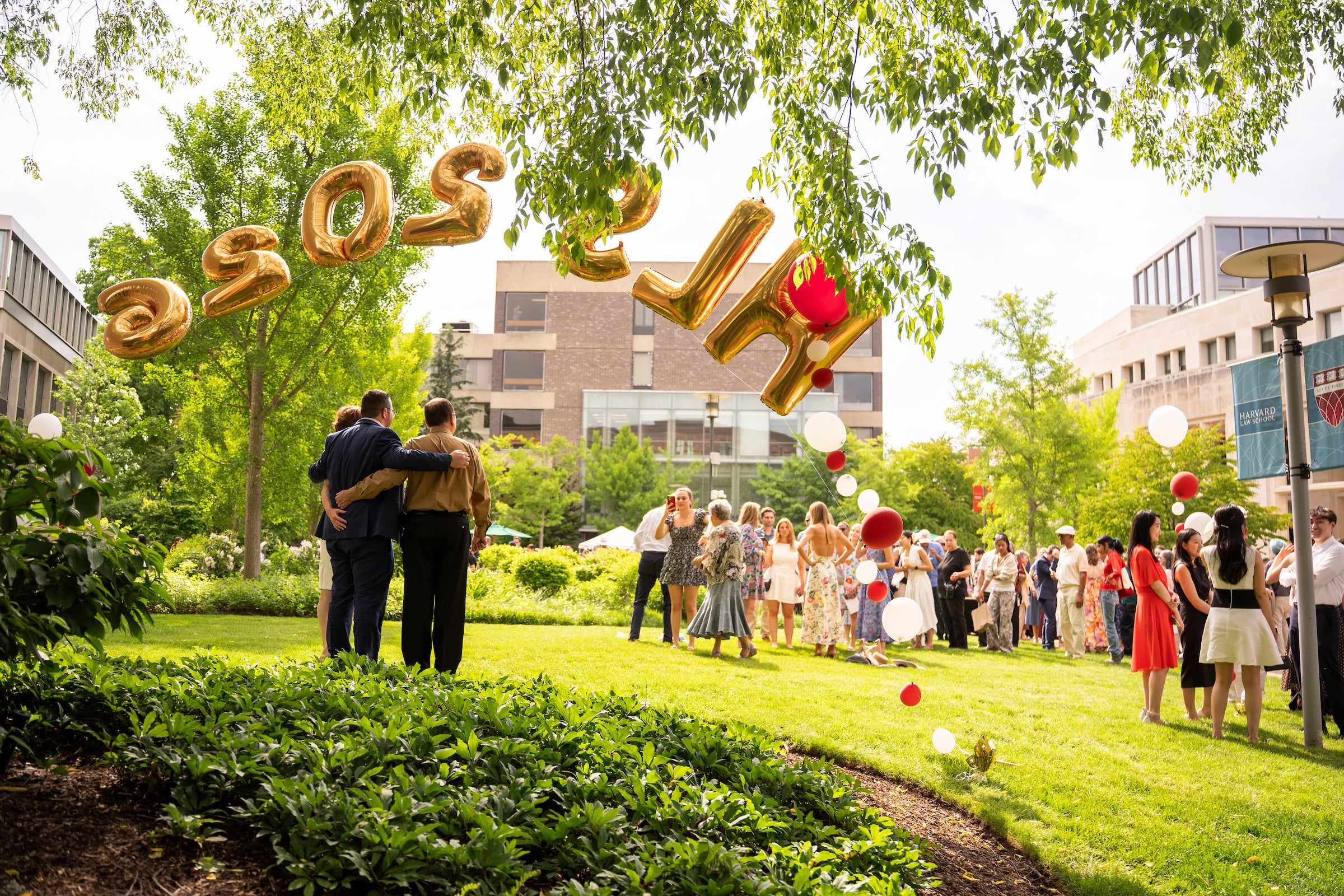 A celebratory gathering outdoors under trees, with three people posing for a photo in front of large gold balloons spelling 