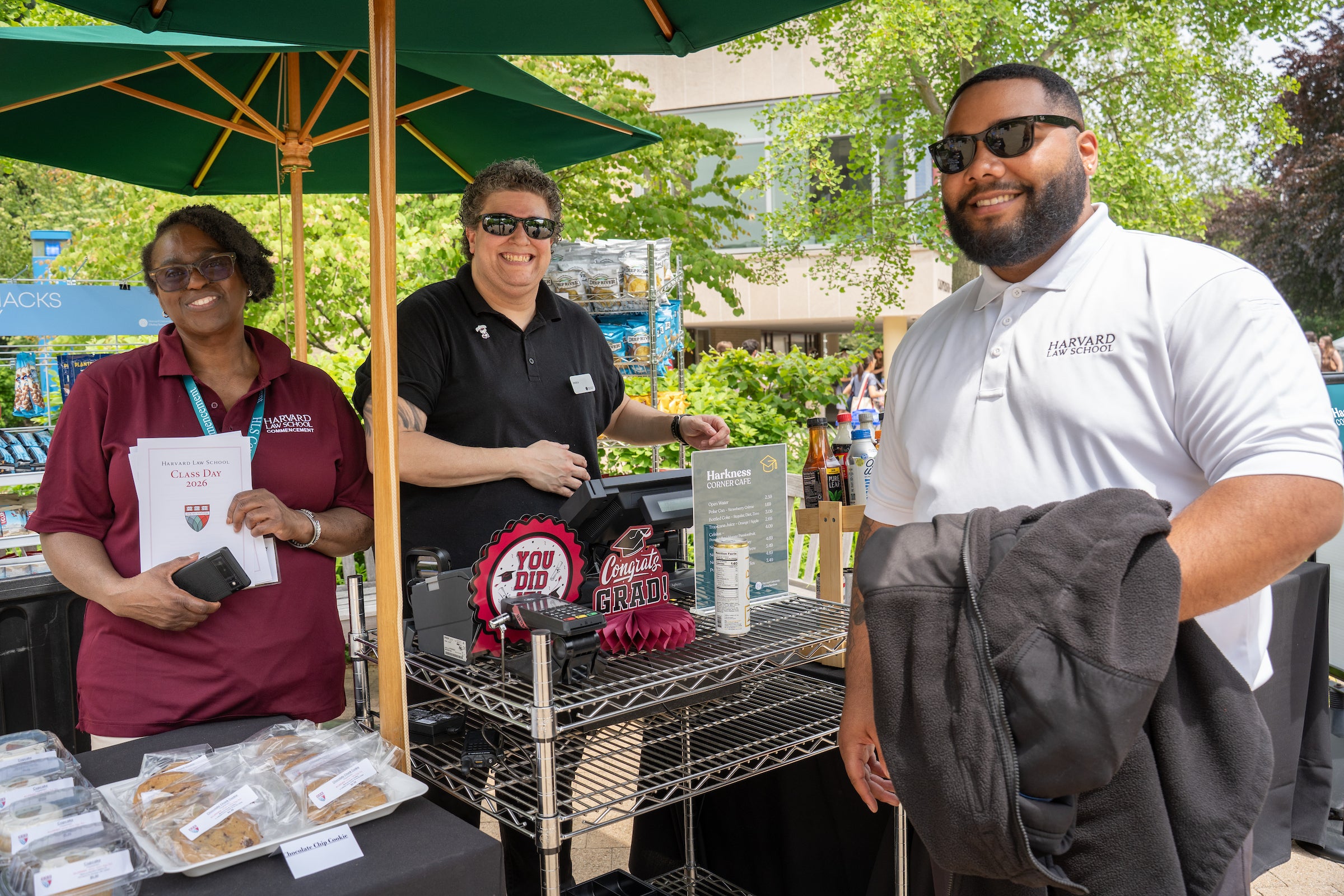 Three individuals stand under a green umbrella at a food stand, with one holding a graduation program and another wearing a Harvard Law School shirt.