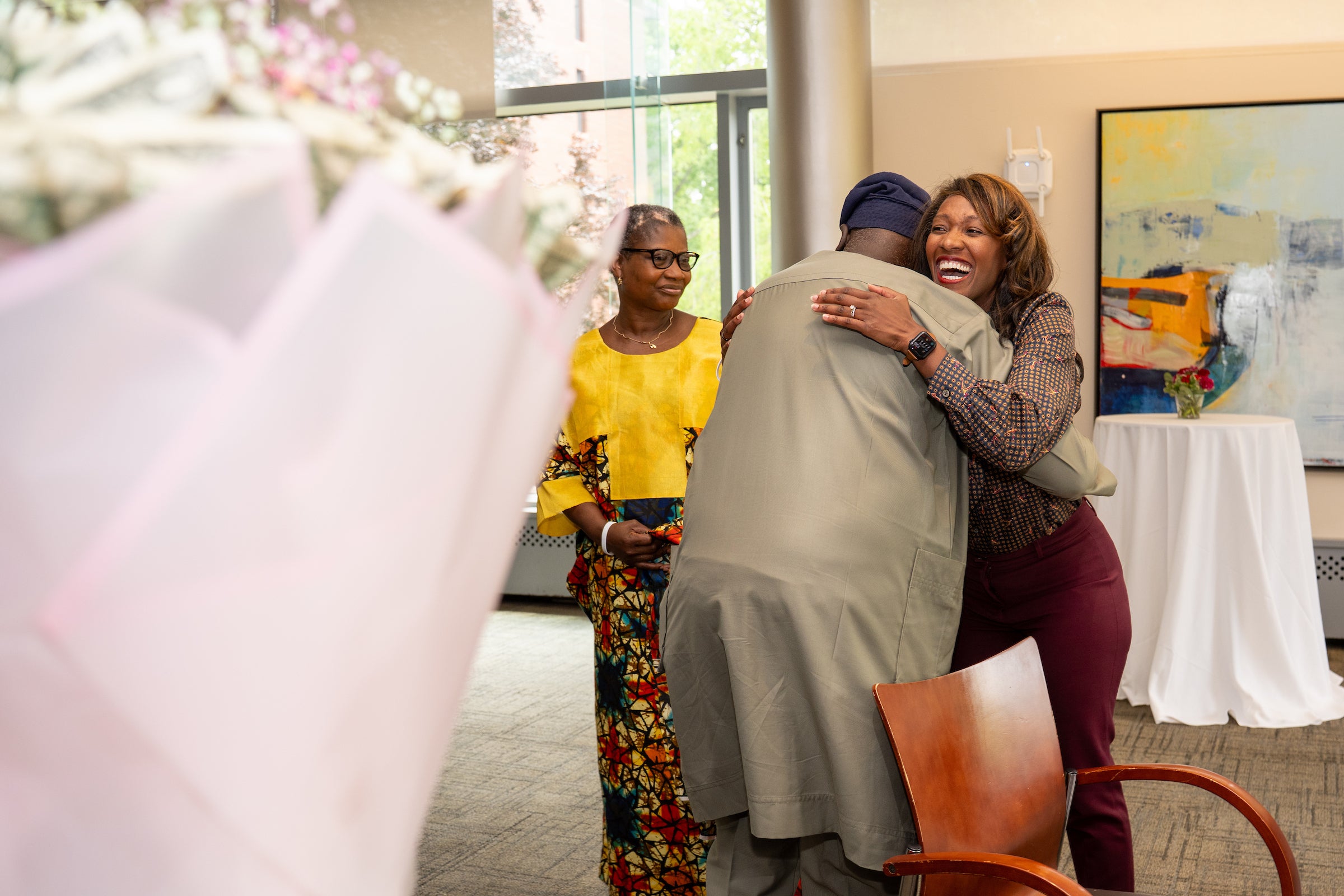 Two individuals embrace warmly in a room, while another person observes nearby, with flowers and artwork visible in the background.