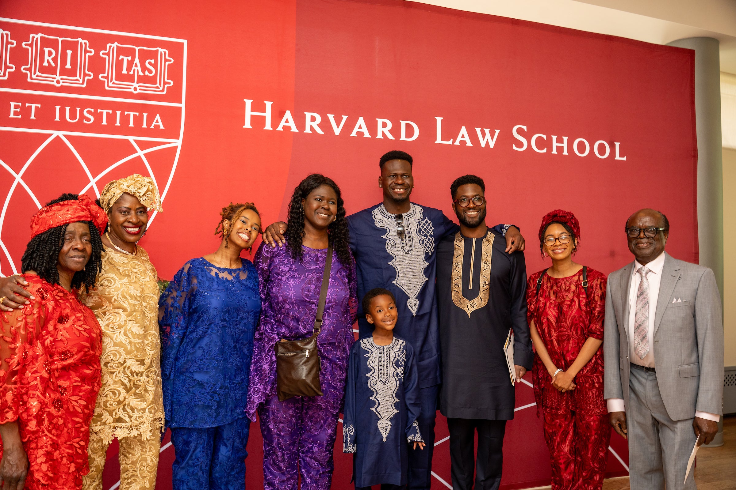 A group of people dressed in vibrant traditional attire pose together in front of a Harvard Law School backdrop.