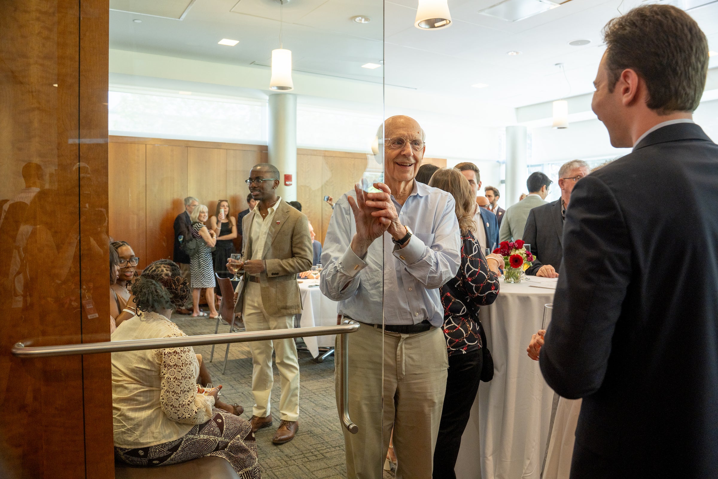 A person holds the door for someone at an indoor event surrounded by tables and other attendees.