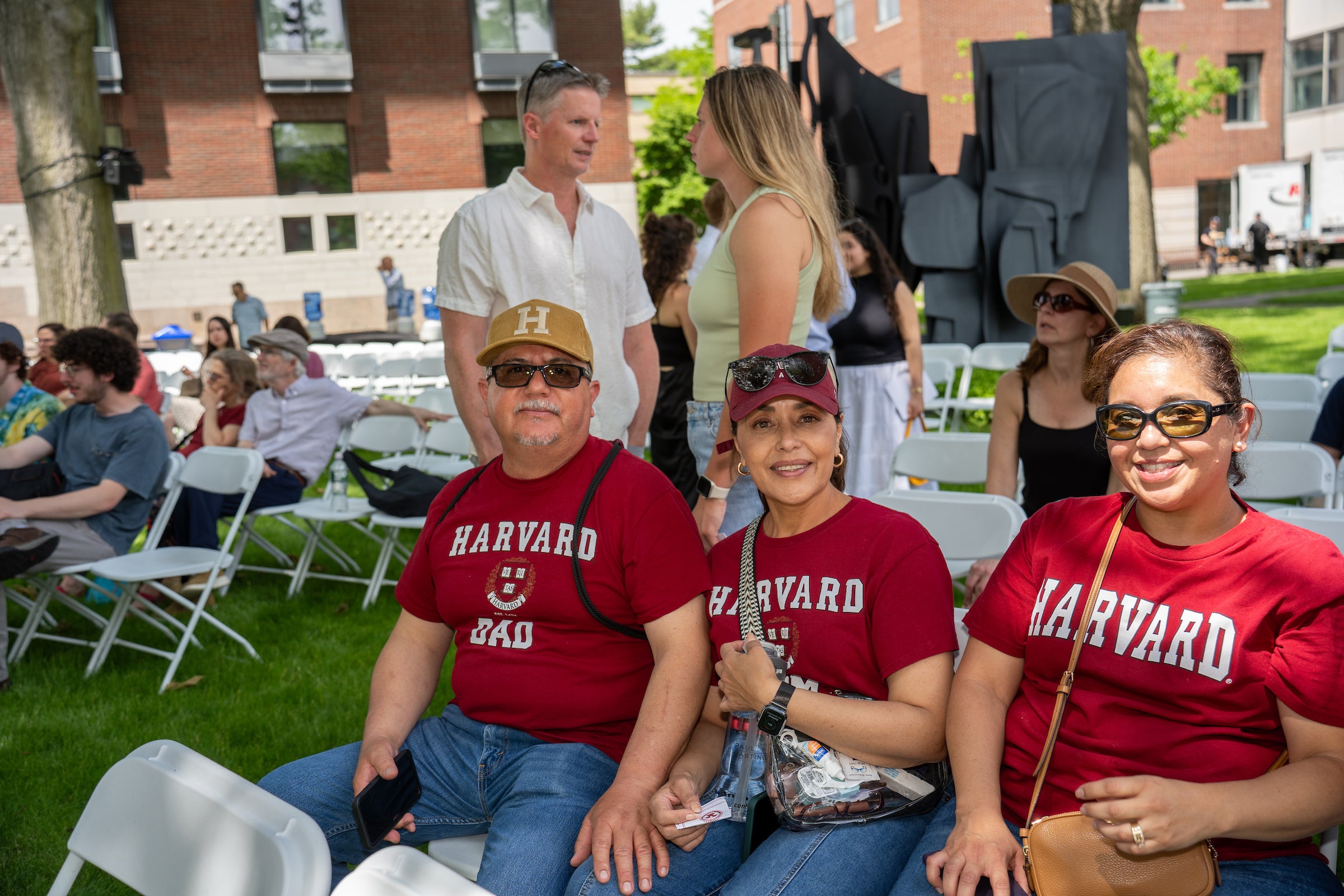 Three individuals wearing Harvard apparel sit together on white chairs at an outdoor event, surrounded by other attendees and a brick building in the background.