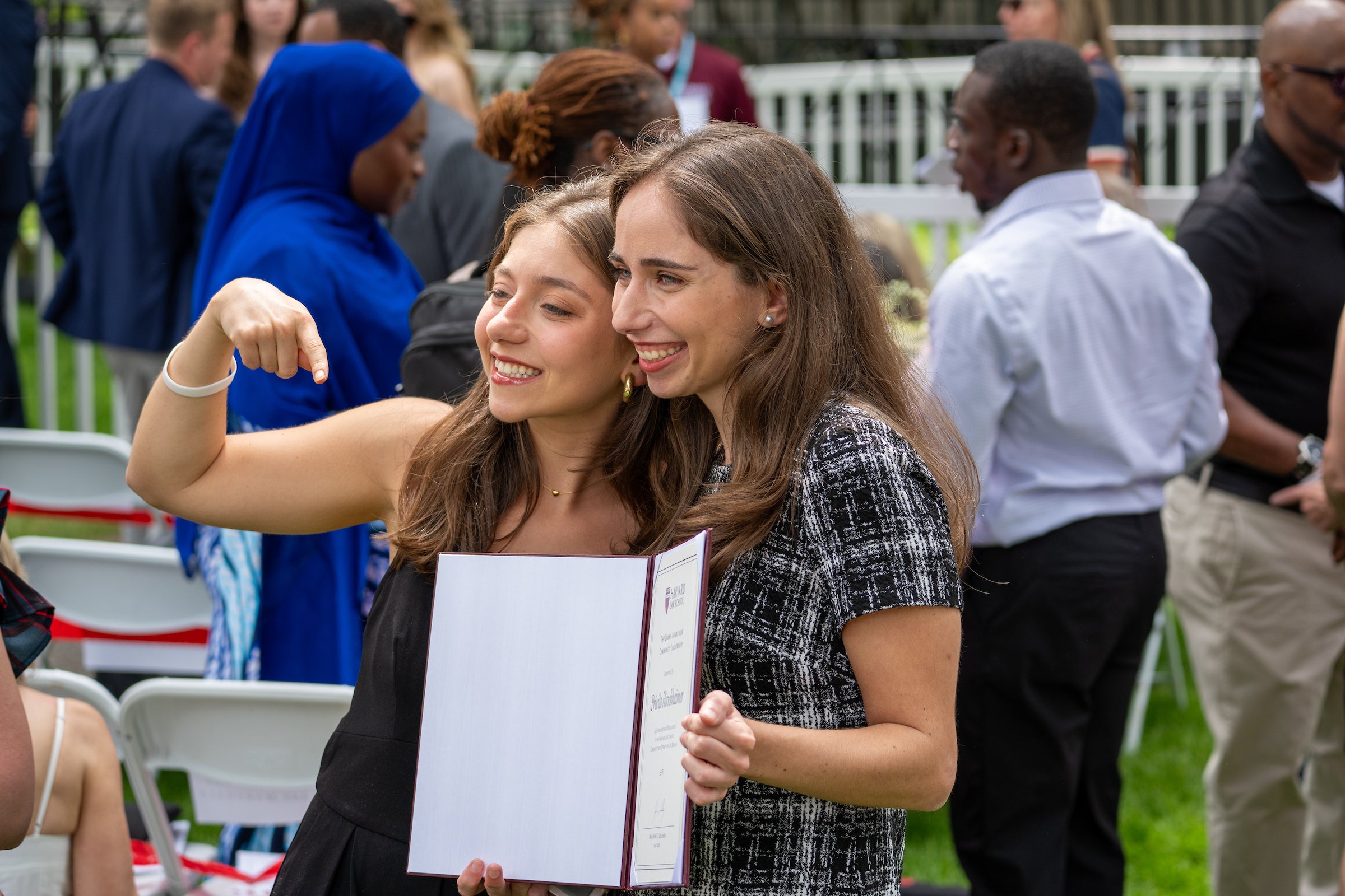 Two individuals smile for a photo at an outdoor event, one pointing at the award that the other is holding.