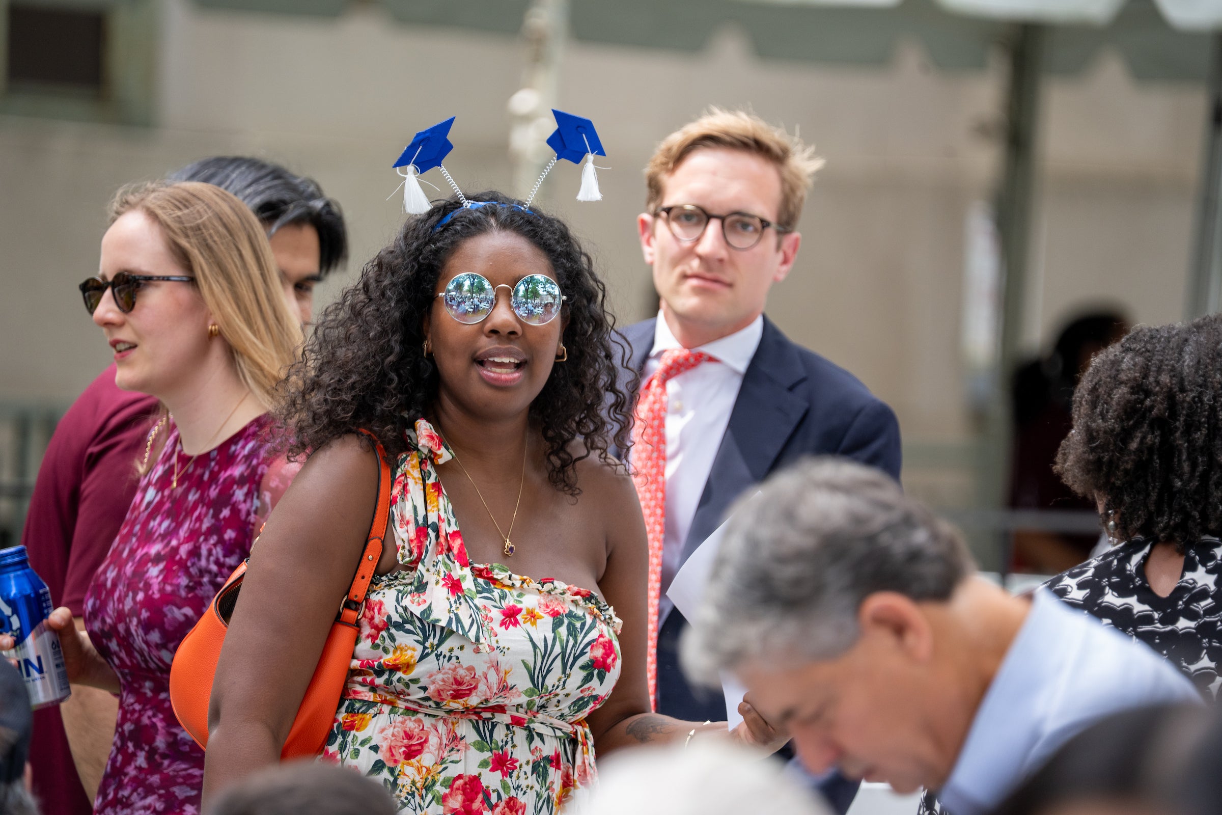A person with curly hair wears a headband decorated with graduation caps and sunglasses while standing in a crowd at an outdoor event.