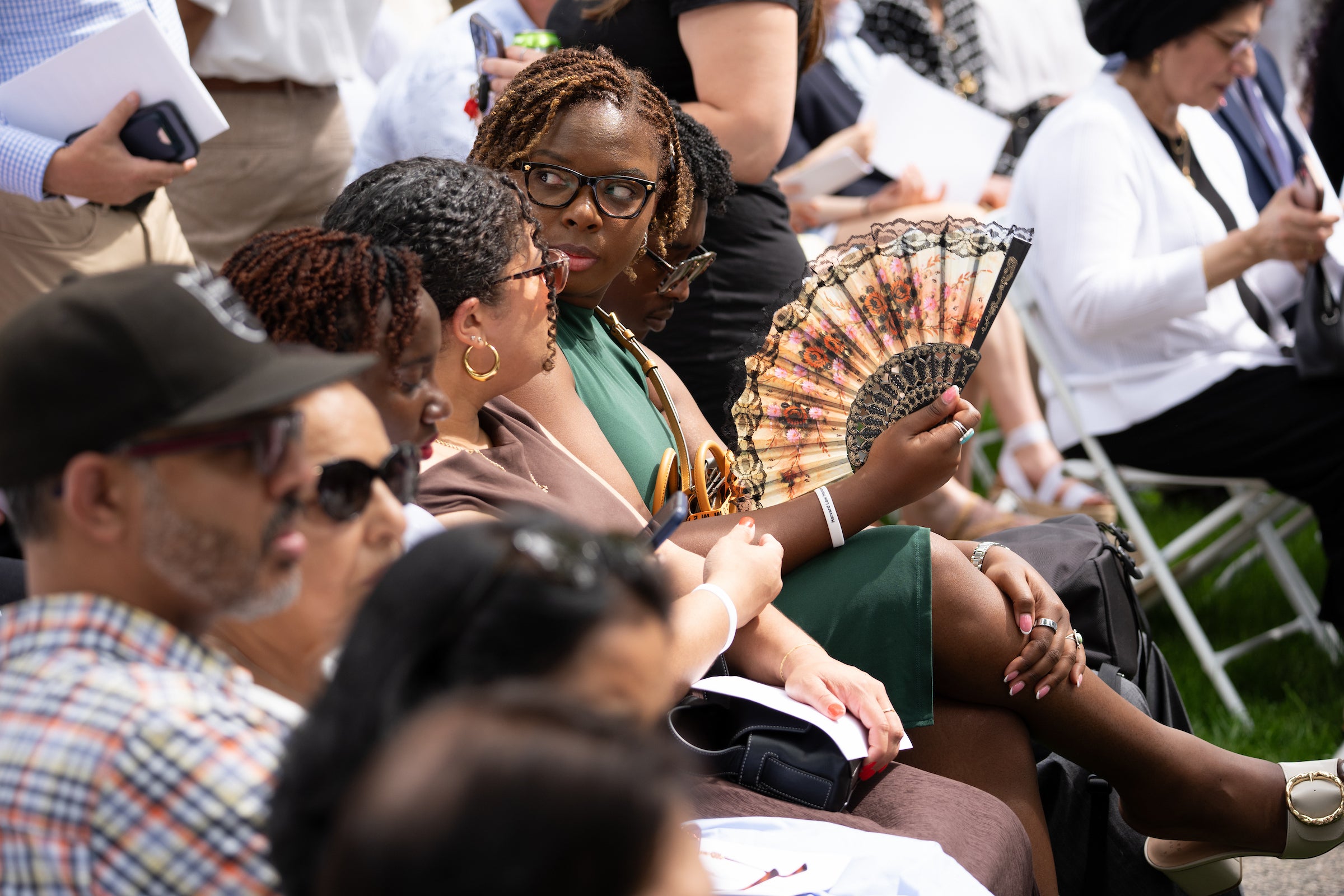 A group of people seated outdoors, with one individual holding a decorative fan and others holding papers or looking around.