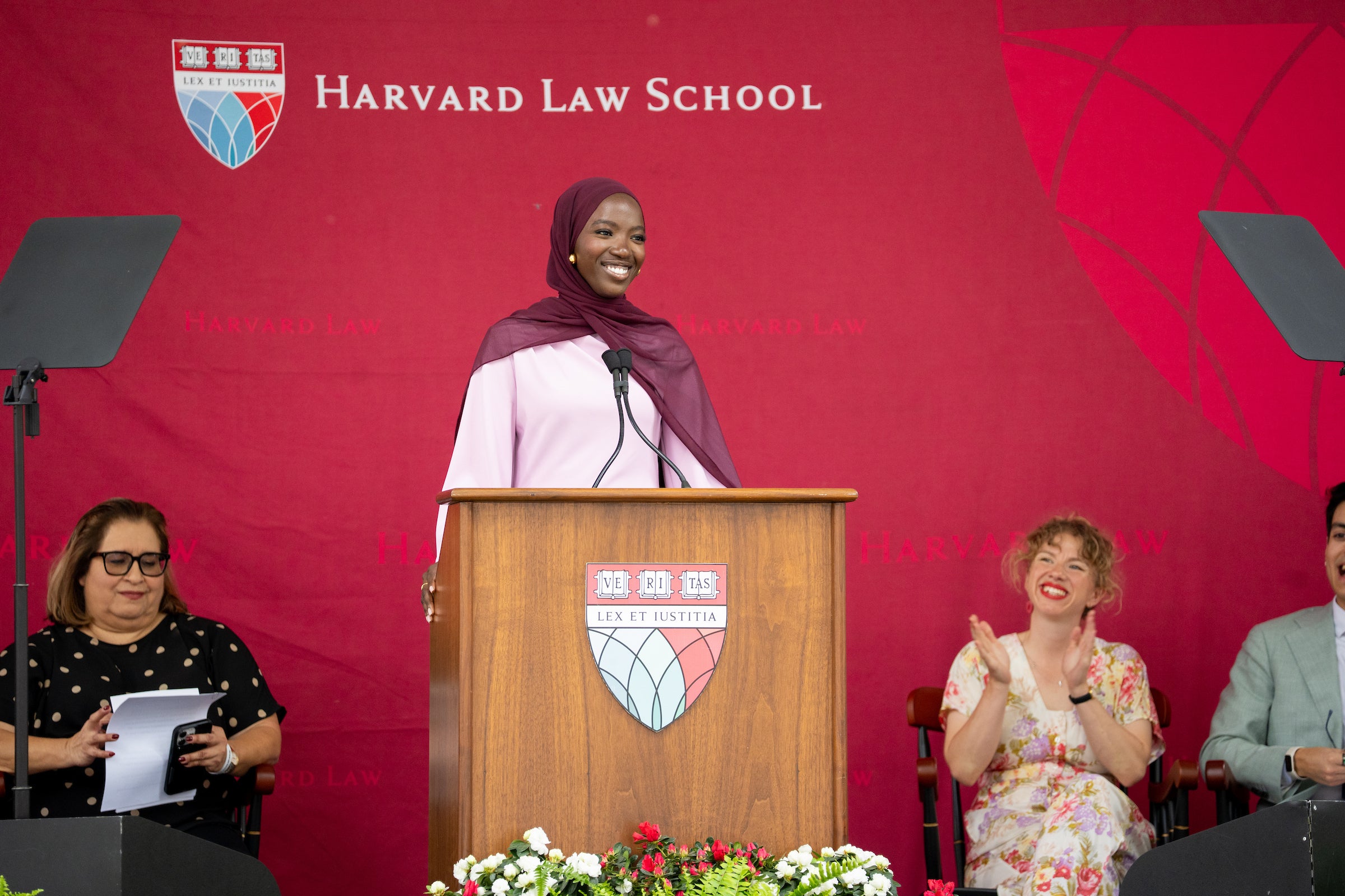 A person in a maroon hijab stands at a wooden podium with the Harvard Law School emblem, smiling while speaking, as others applaud and watch from the side.