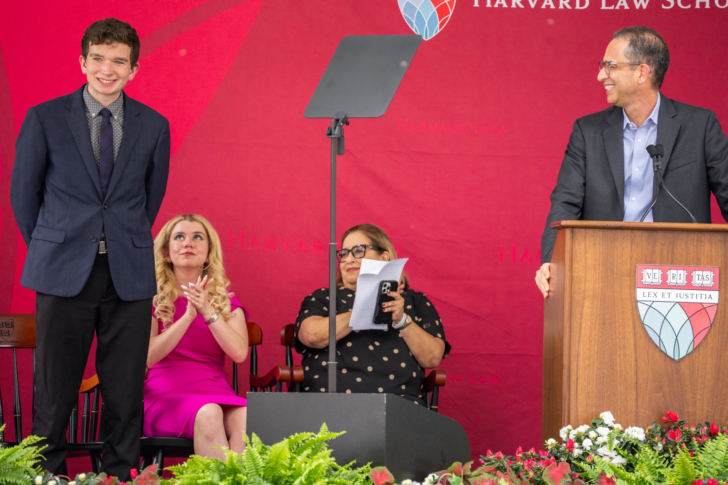 Dean Goldberg speaks at a podium on a stage, while others seated nearby applaud a graduate, who is standing.