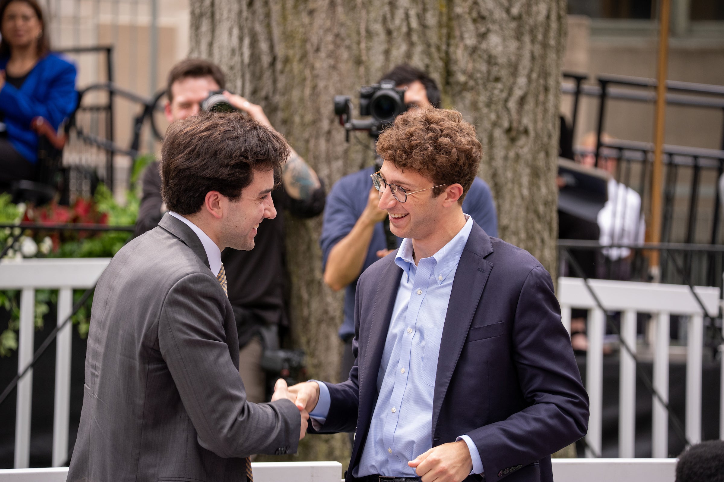 Two individuals in suits shake hands outdoors, smiling, with photographers and a tree visible in the background.