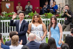 Three individuals wave to an audience during an outdoor ceremony, with a photographer capturing the moment in the background.