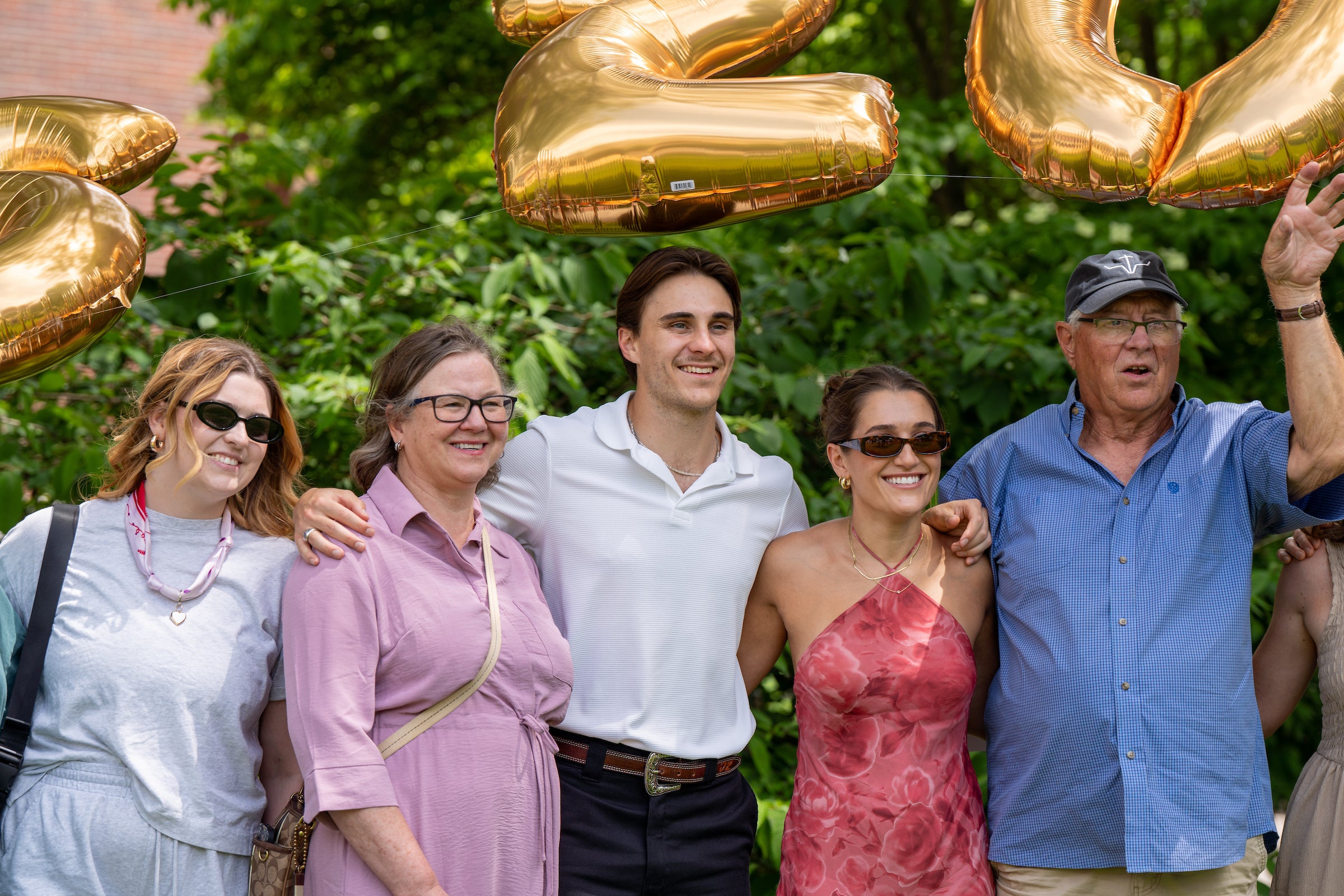 A group of five people pose outdoors under large golden balloons, smiling for the camera.