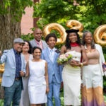 A graduate in a cap holding a bouquet of flowers stands with family and friends outdoors, smiling in front of large gold “2026” balloons.