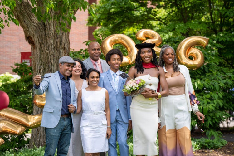 A graduate in a cap holding a bouquet of flowers stands with family and friends outdoors, smiling in front of large gold “2026” balloons.