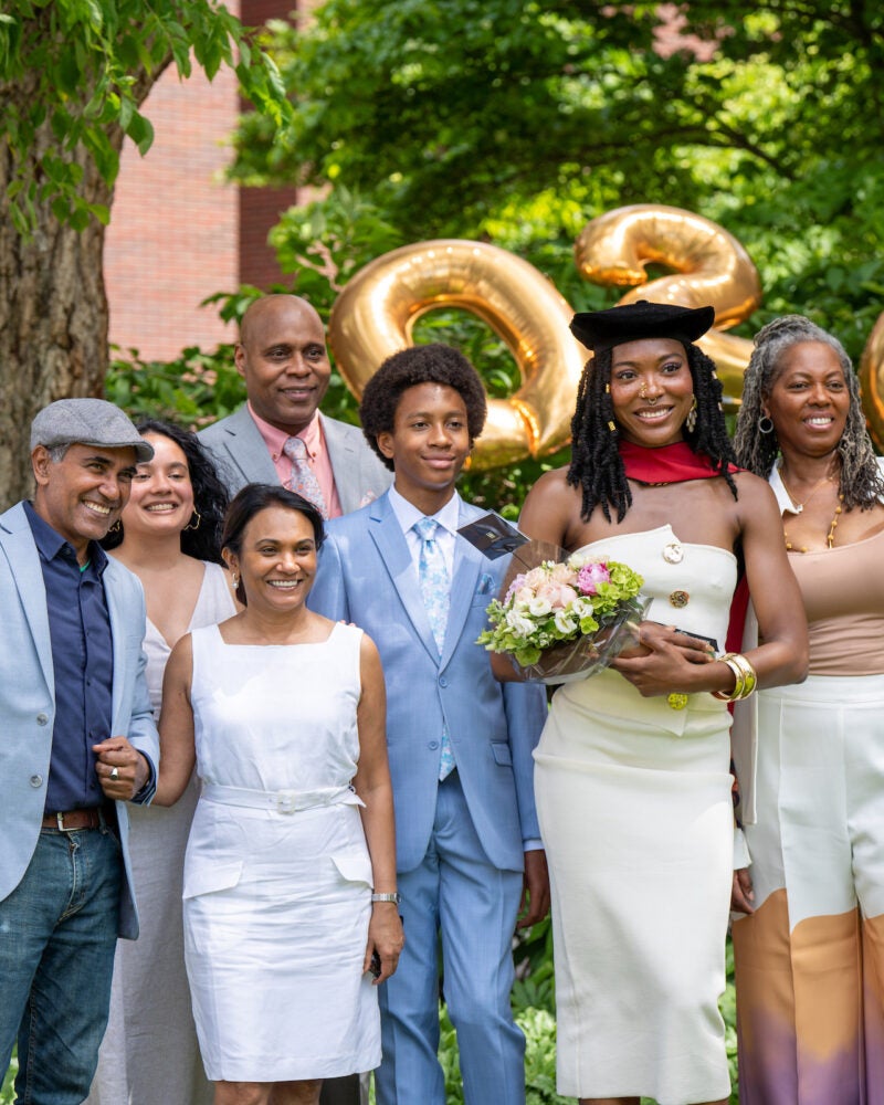 A graduate in a cap holding a bouquet of flowers stands with family and friends outdoors, smiling in front of large gold “2026” balloons.