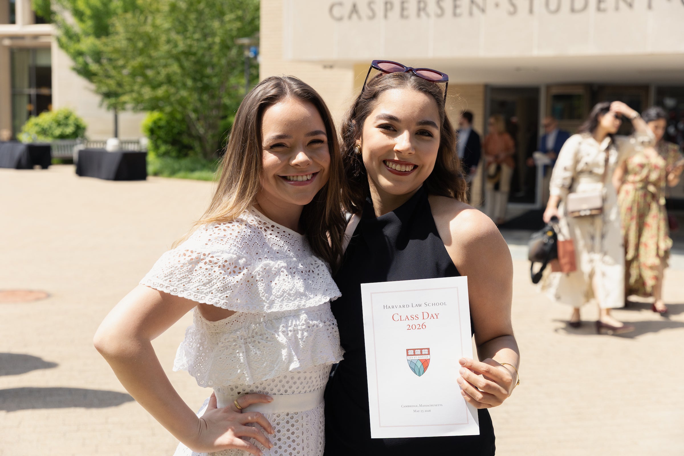 Two individuals smile for the camera at Harvard Law School’s Class Day 2026, with one holding a program and sunglasses perched on their head.