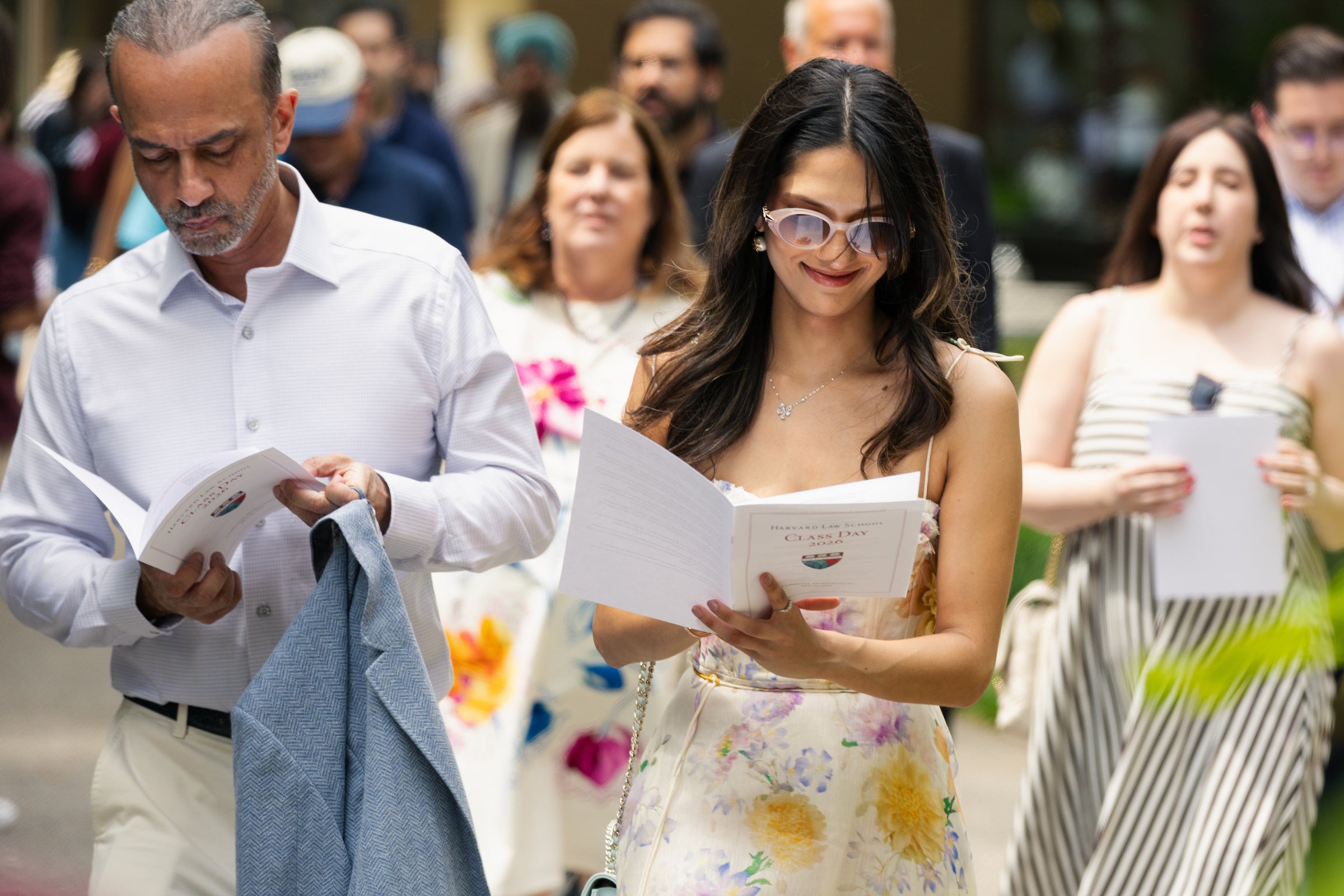 A group of people walk outdoors, holding event programs, with one person smiling while wearing sunglasses and a floral dress.