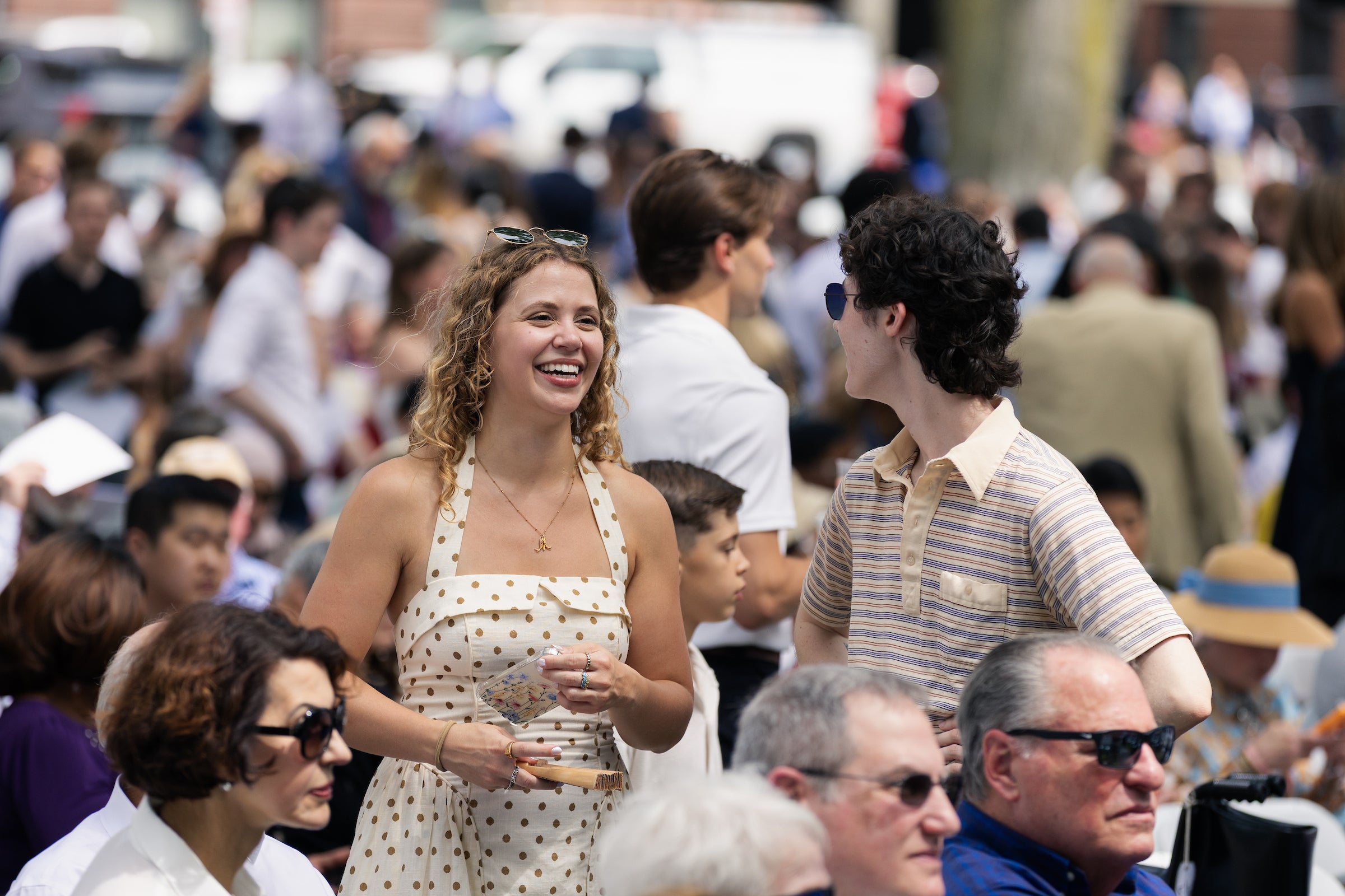 A smiling person in a polka-dot dress holds a drink while standing in a crowded outdoor gathering, surrounded by others enjoying the event.
