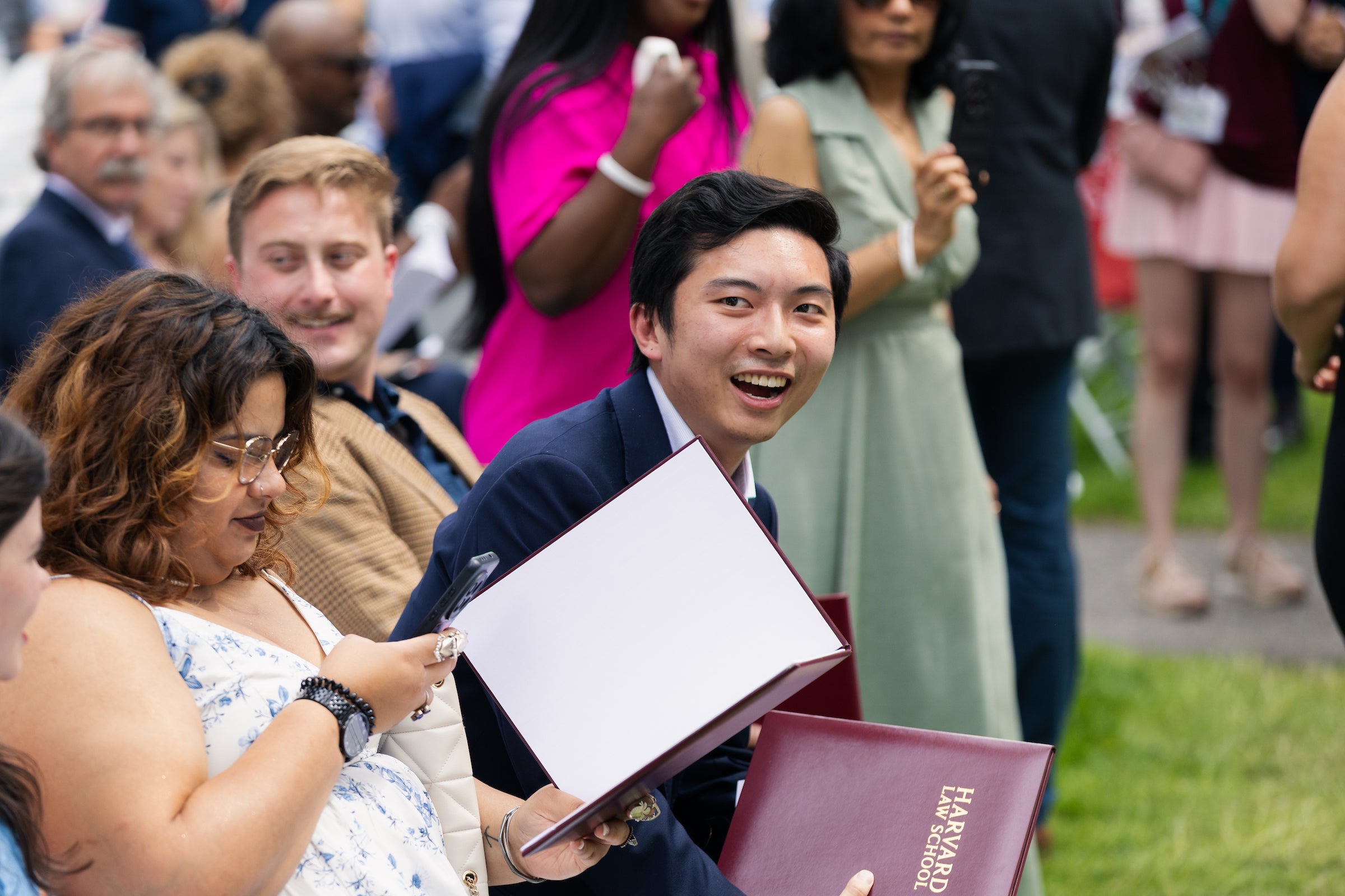 A person in a suit holds a Harvard Law School folder and smiles broadly while seated among an audience at an outdoor event.