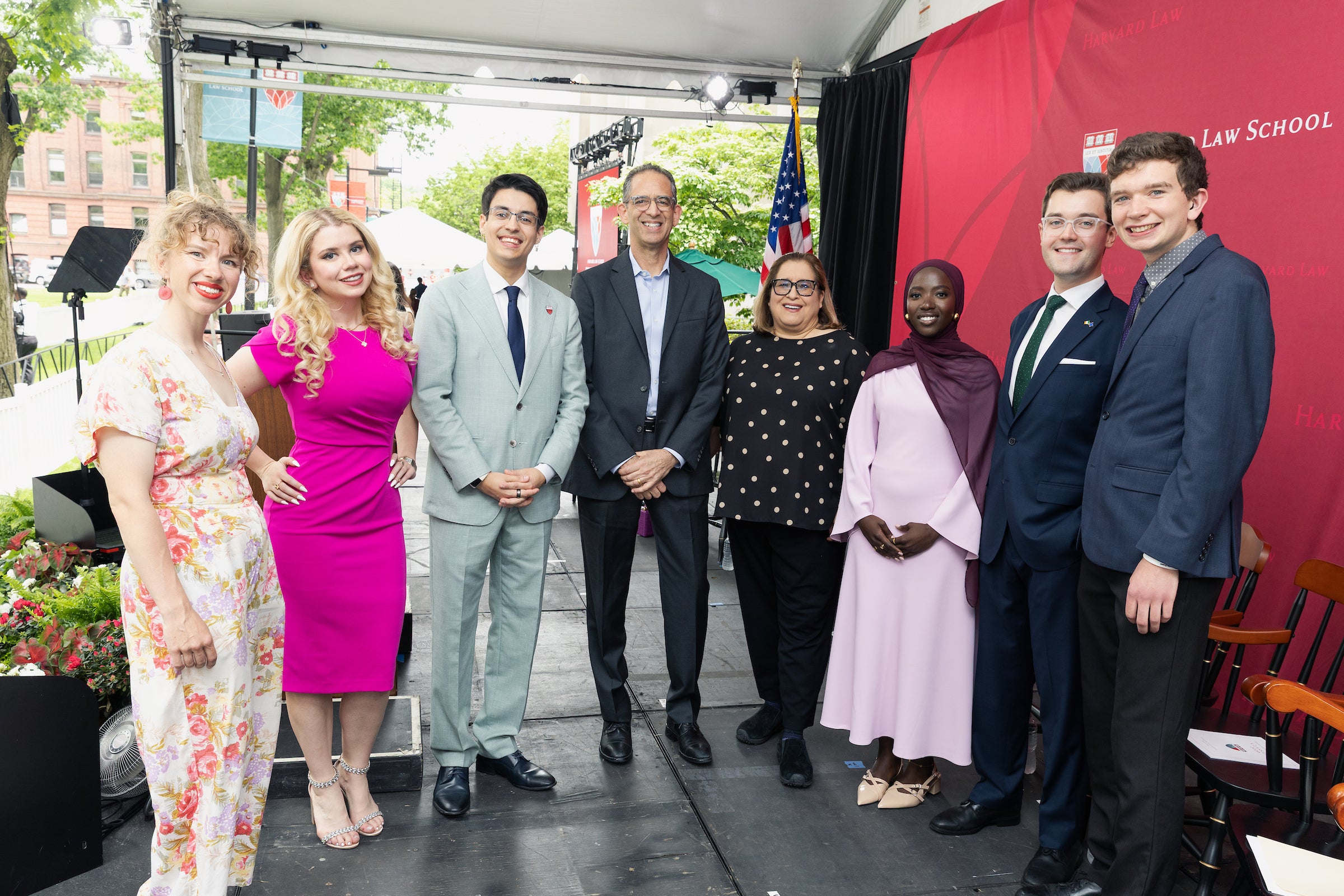 A group of eight individuals poses together on a stage at an outdoor event, with a Harvard Law School banner visible in the background.