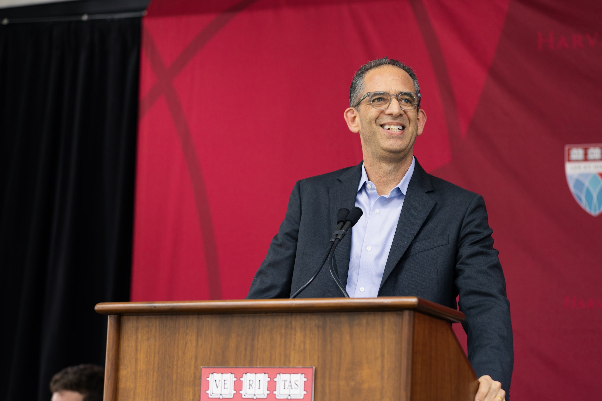 Dean Goldberg stands at a wooden podium with a microphone, smiling, against a red backdrop.