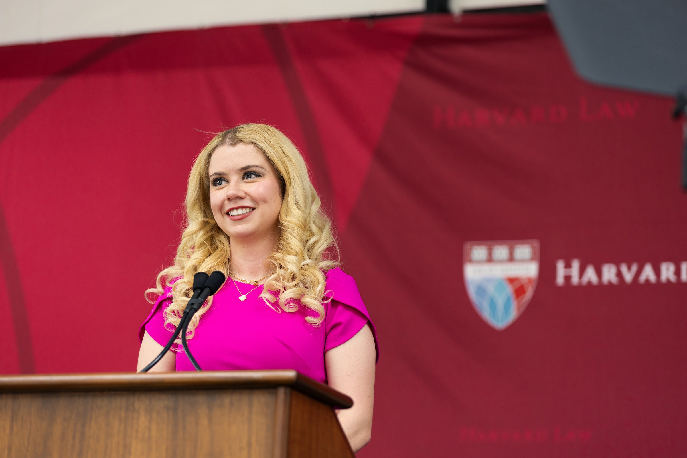 A person with long blonde hair stands at a podium in front of a red backdrop, smiling while speaking into microphones.