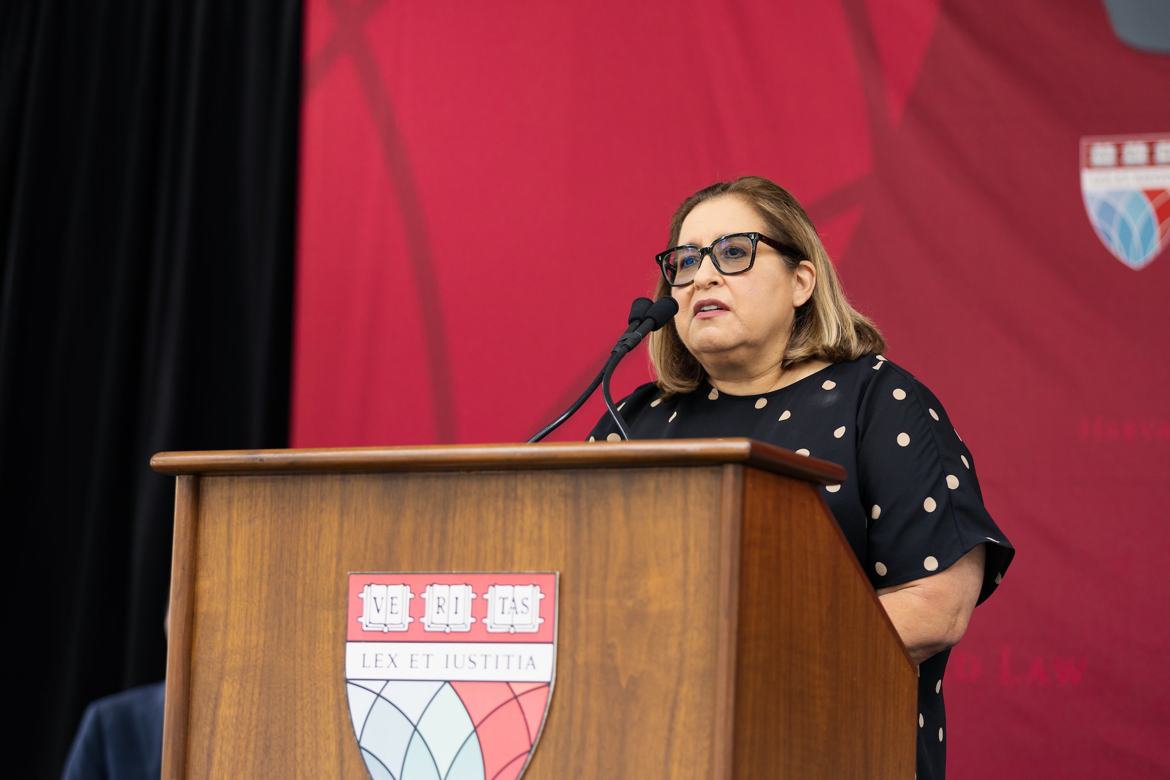 A person with glasses speaks at a wooden podium adorned with the Harvard Law shield, standing before a red backdrop.