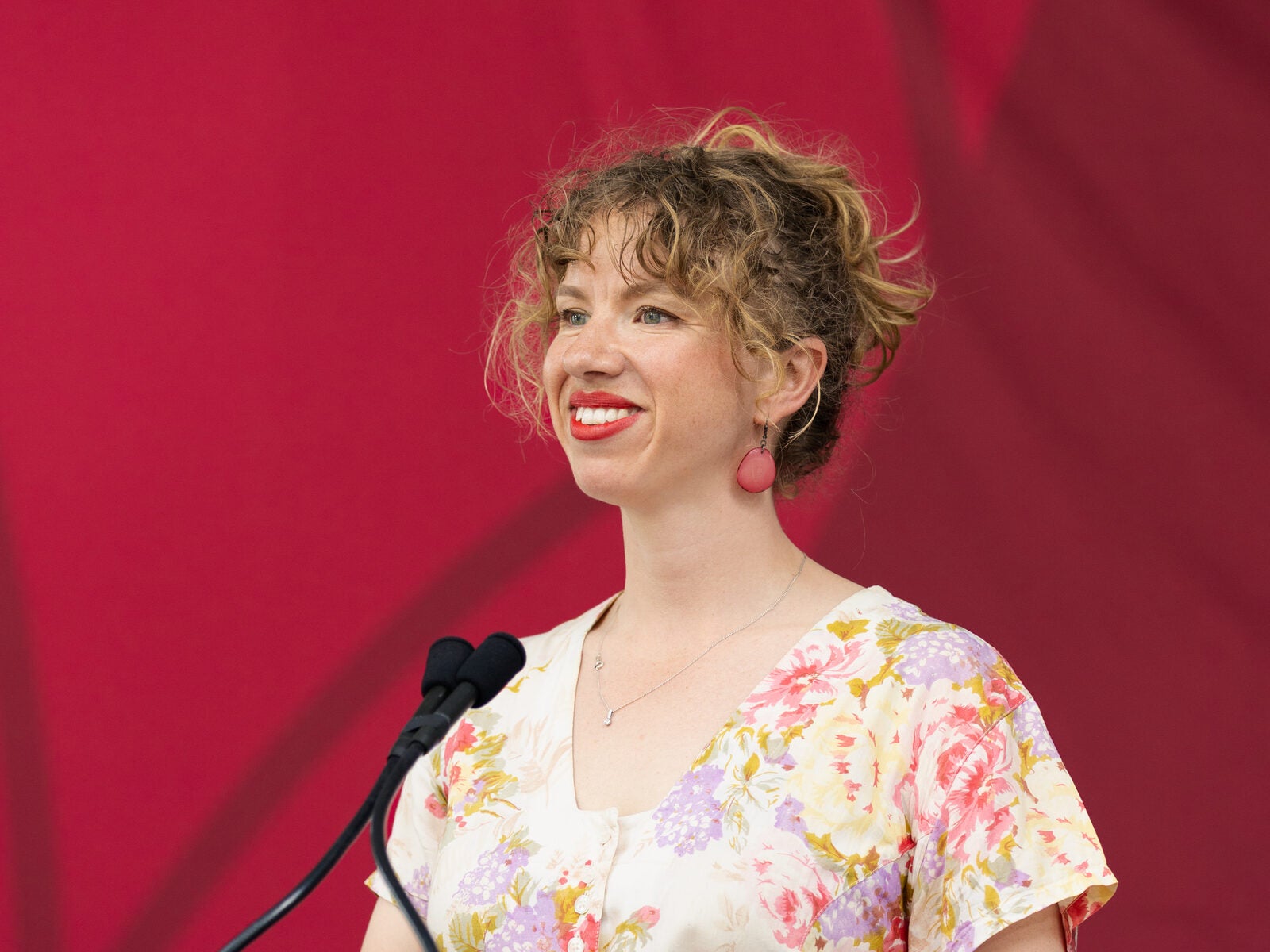 A person with curly hair and a floral dress stands at a podium, smiling while speaking into a microphone against a red backdrop.