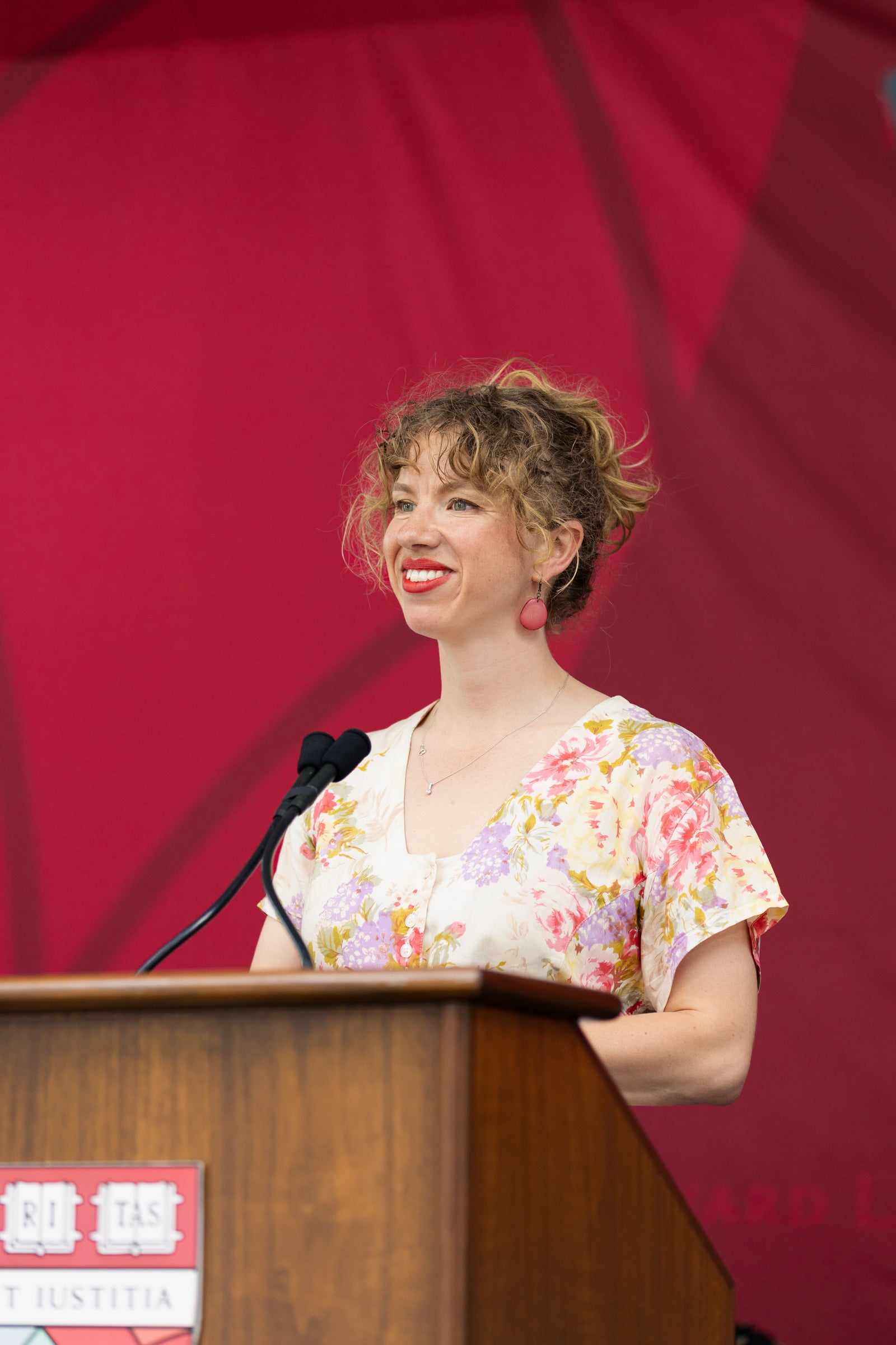 A person with curly hair and a floral dress stands at a podium, smiling while speaking into a microphone against a red backdrop.