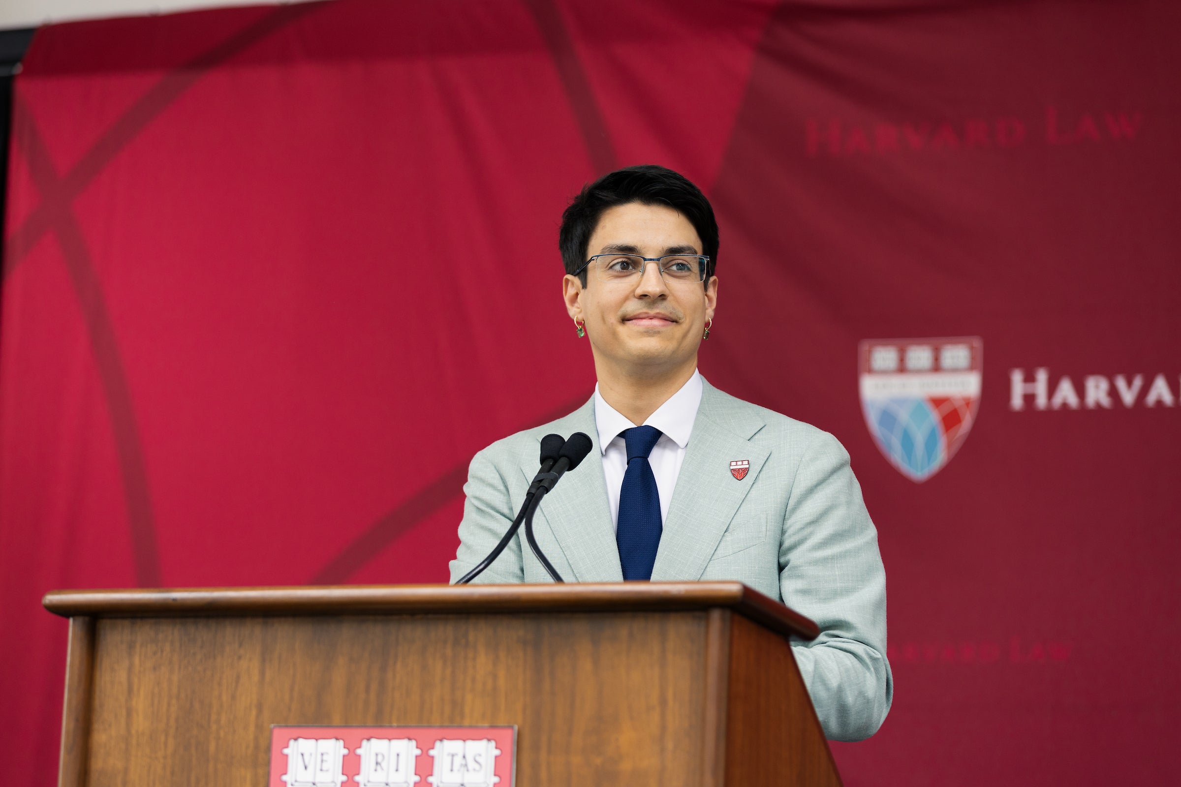 A person in a light gray suit and blue tie stands at a wooden podium, speaking into microphones in front of a red backdrop.
