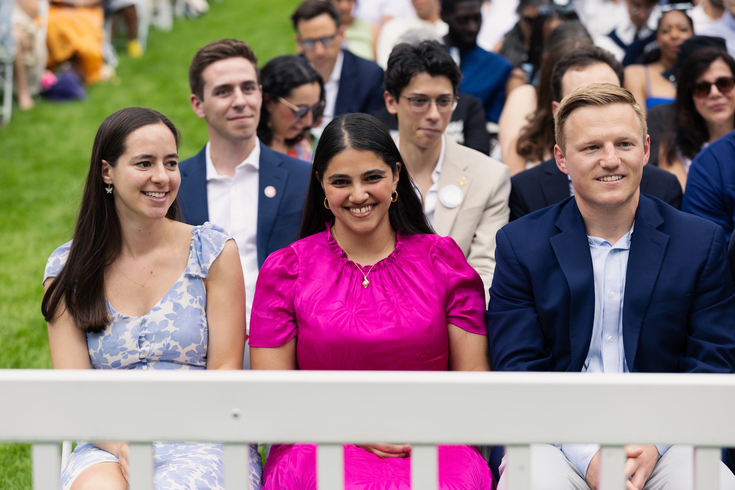 A group of people, including a person in a bright pink dress, are seated outdoors at an event, smiling and looking toward the camera.